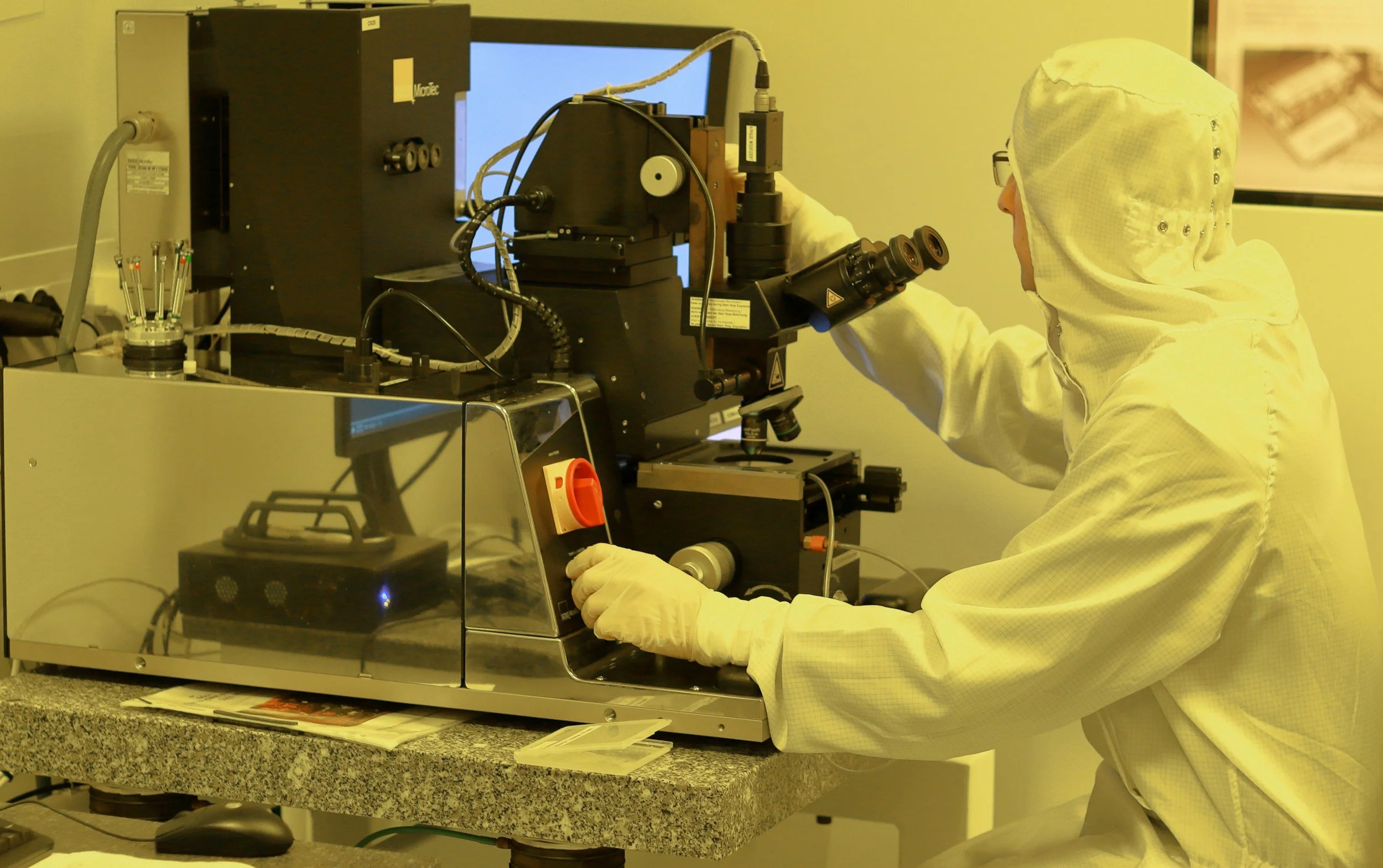 A scientist in a yellow lab coat and safety goggles working with a microscope and scientific equipment in a laboratory setting.