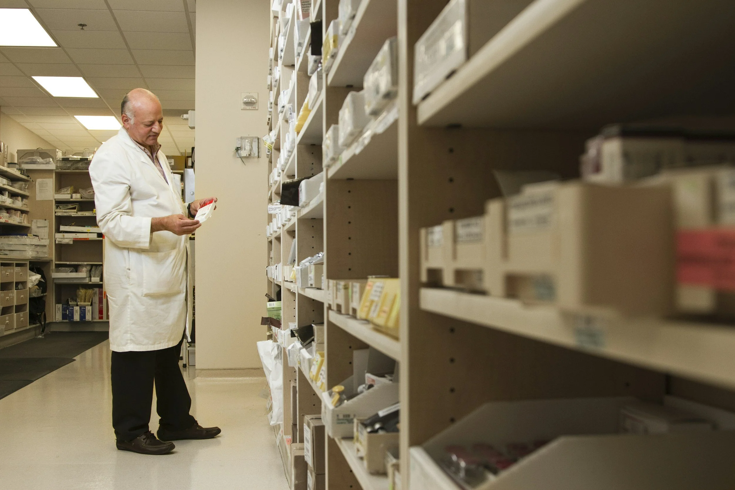 A man in a white lab coat shopping in a pharmacy aisle, looking at a product in his hand.