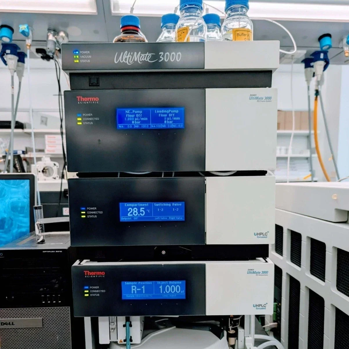 Laboratory equipment with three stacked modules, each displaying digital readouts: the top has bottles of liquids, the middle shows pump settings, and the bottom shows temperature and sample volume details.