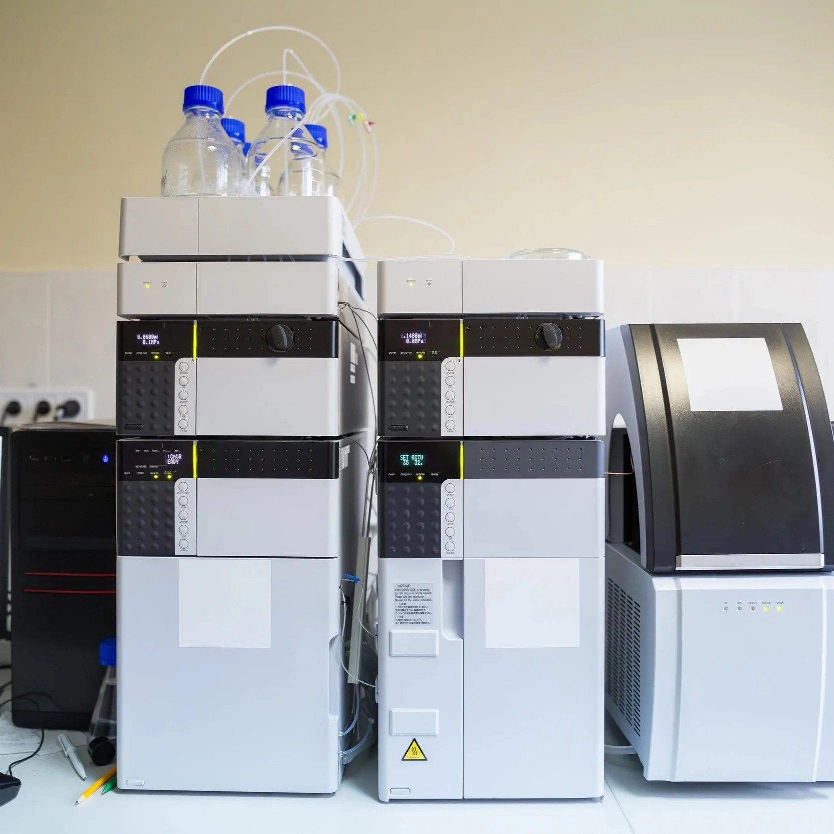 Laboratory setup with multiple scientific instruments stacked on top of each other, including bottles with tubing, placed on a white counter against a yellow wall.
