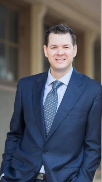 A man in a navy suit and blue tie smiling outdoors in front of a building.