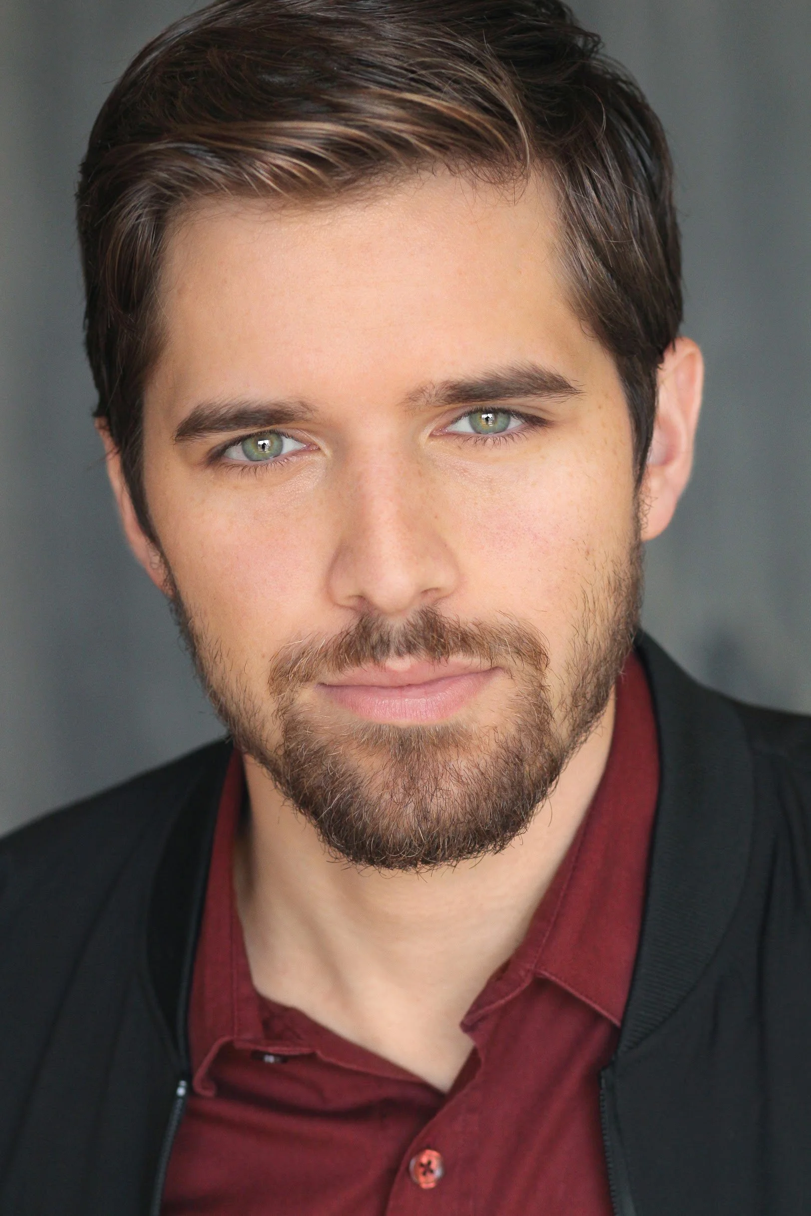 A man with light skin, green eyes, dark brown hair, and a beard wearing a black jacket over a maroon shirt with a black strap visible.