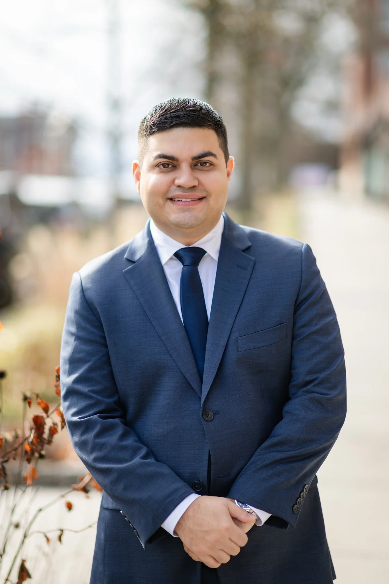 A man in a blue suit with a white shirt and blue tie standing outdoors, smiling, with trees and buildings in the background.