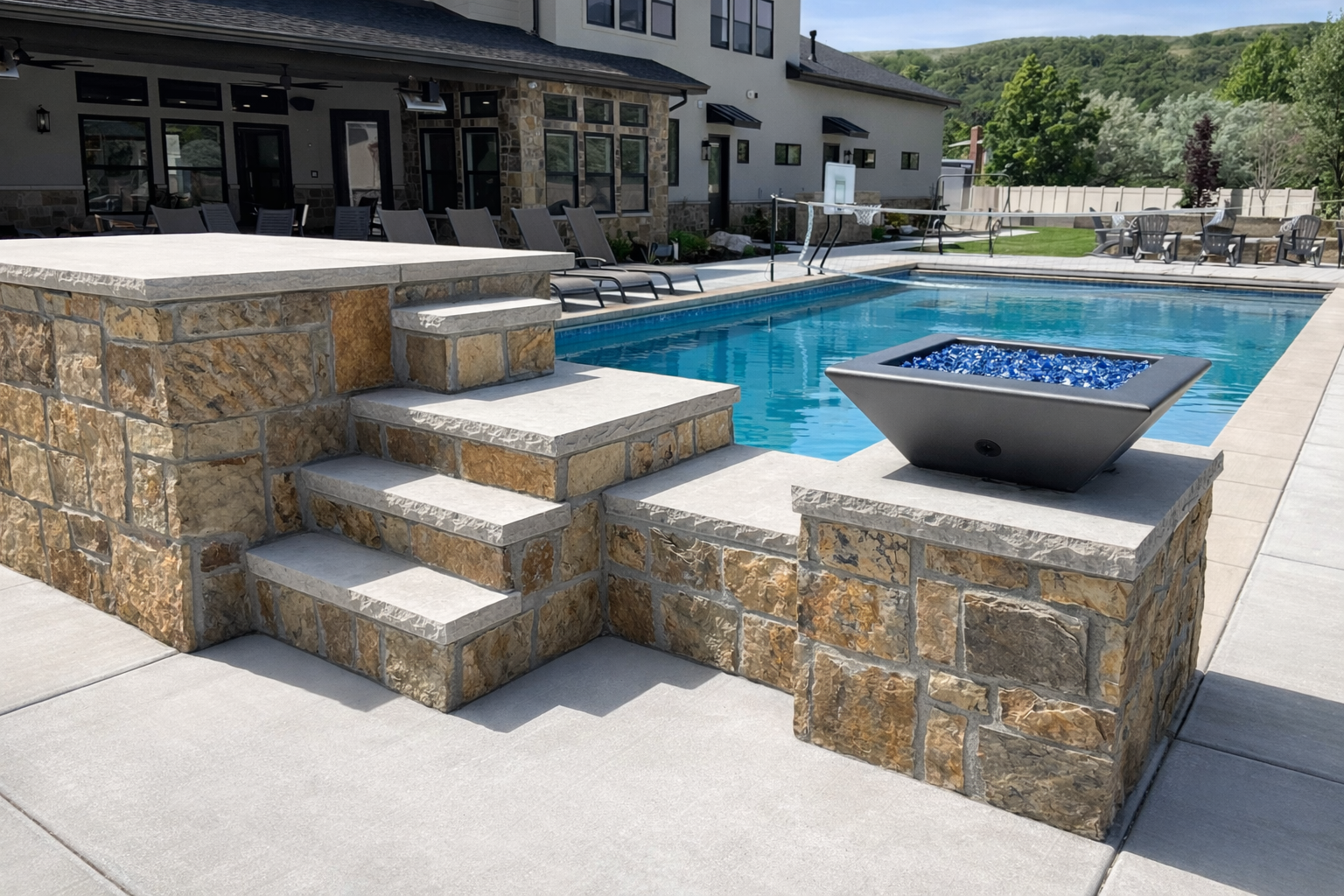 A backyard swimming pool with stone steps and a modern fire bowl, surrounded by patio furniture and a house with large windows, set against a background of green hills and trees.