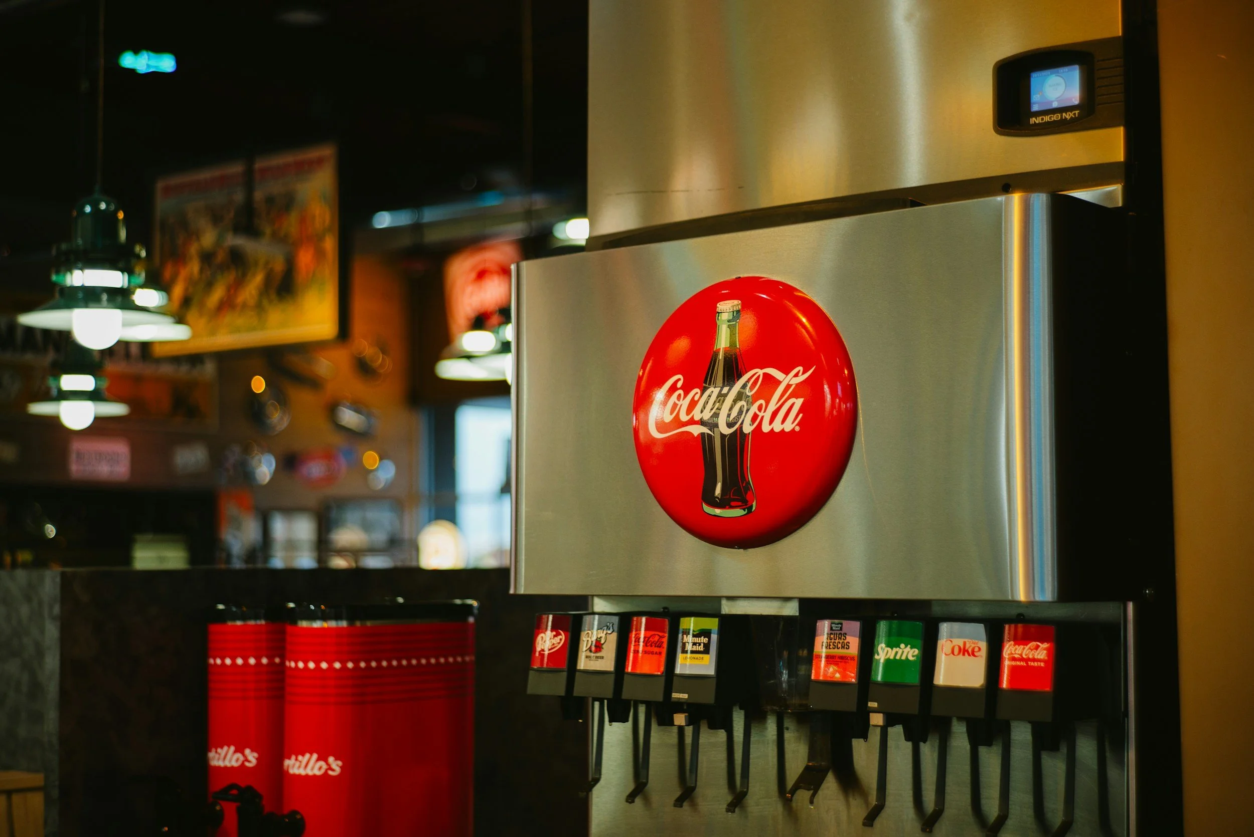 A soda fountain machine with Coca-Cola logo dispensing options in a restaurant or cafe setting with blurred background and warm lighting.