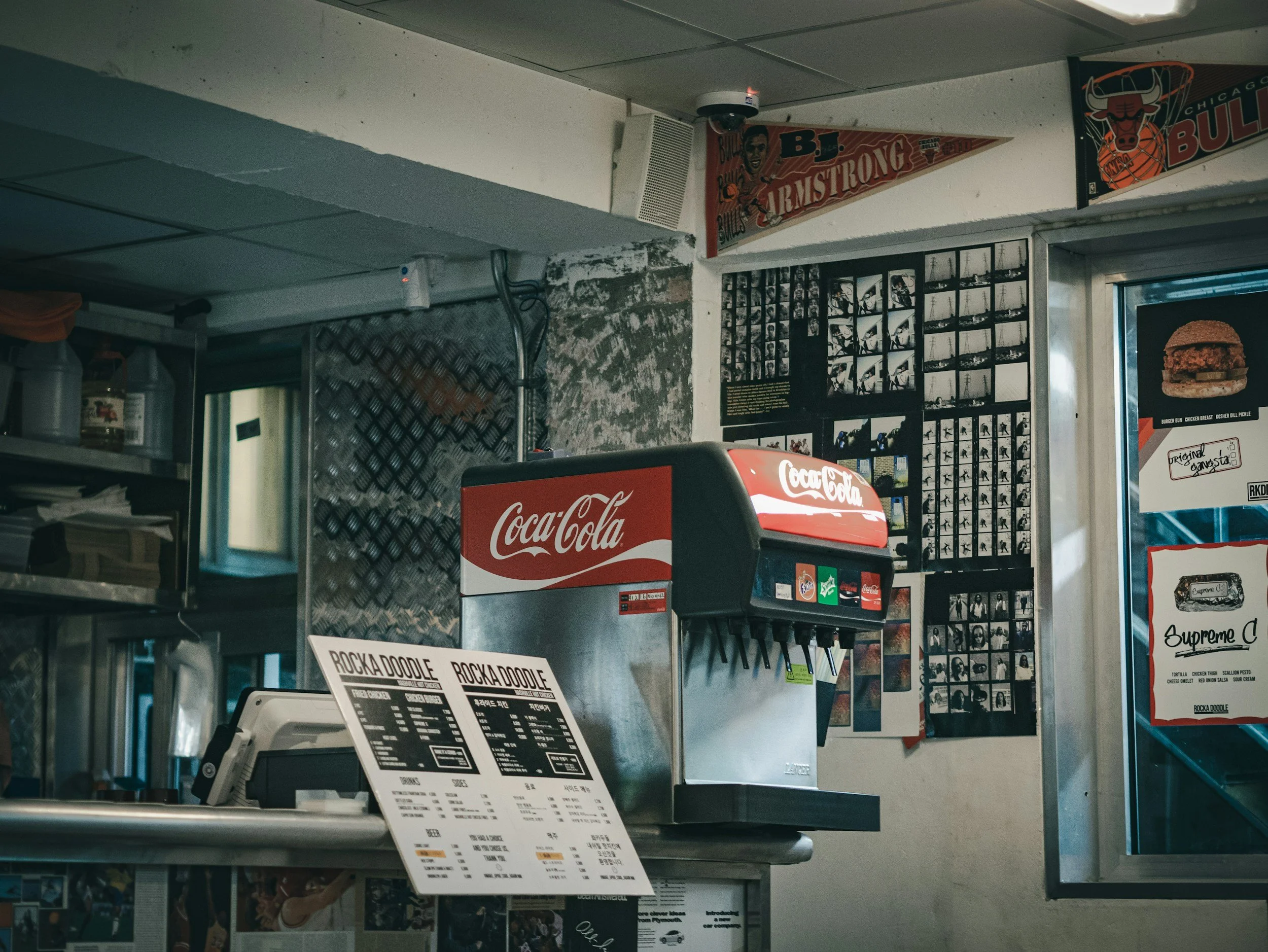 Interior of a fast food restaurant featuring a Coca-Cola soda fountain, menu boards, and decorative posters on the wall.