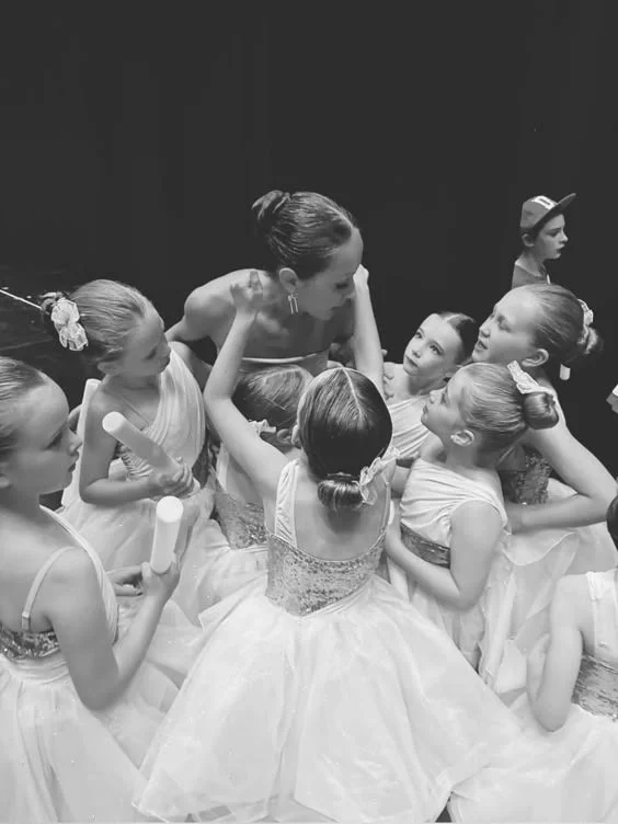 Ballet instructor with young students backstage before a performance, all in tutus.