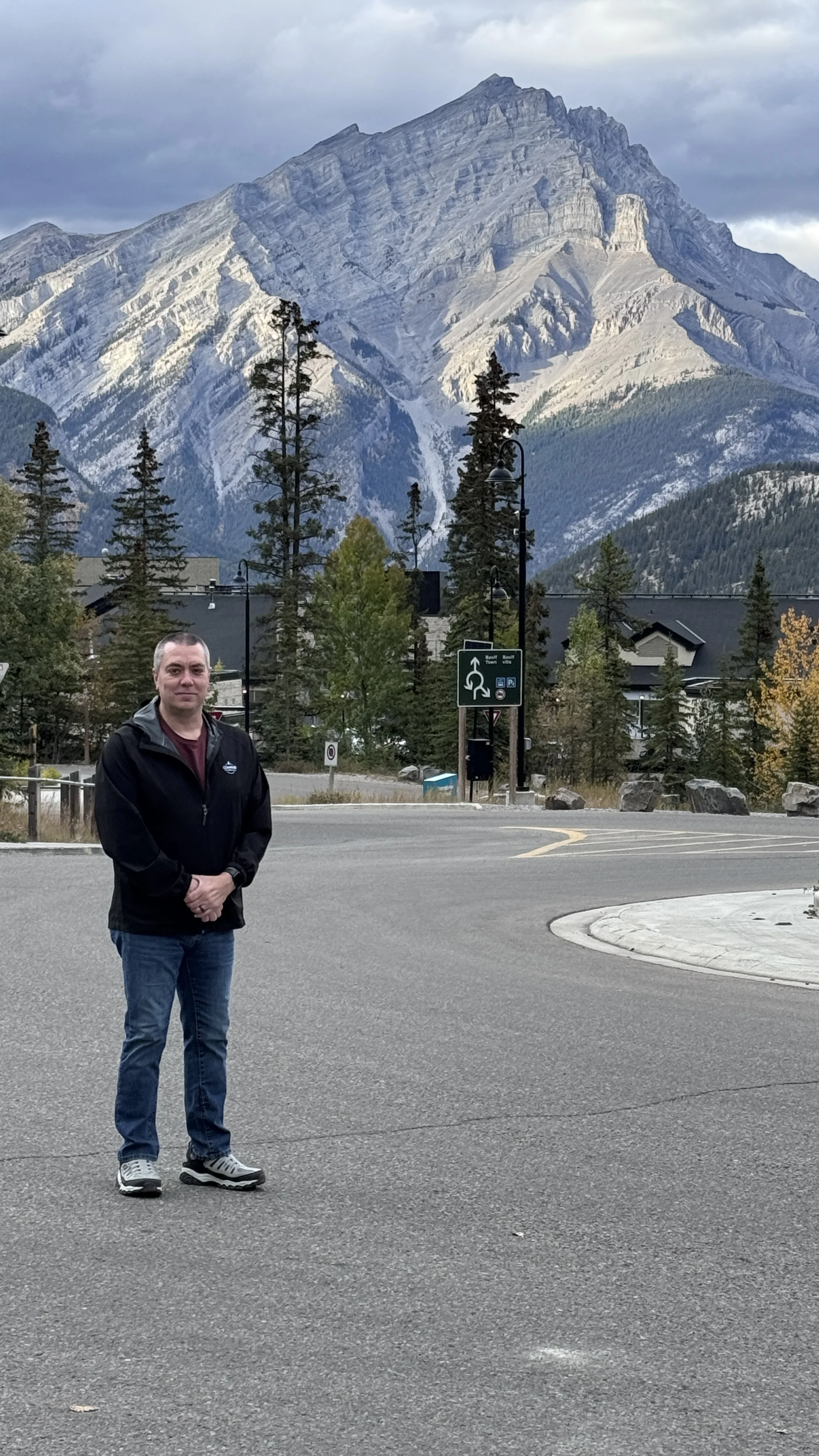 A man in a black jacket and jeans standing in a parking lot with mountains, trees, and buildings in the background.