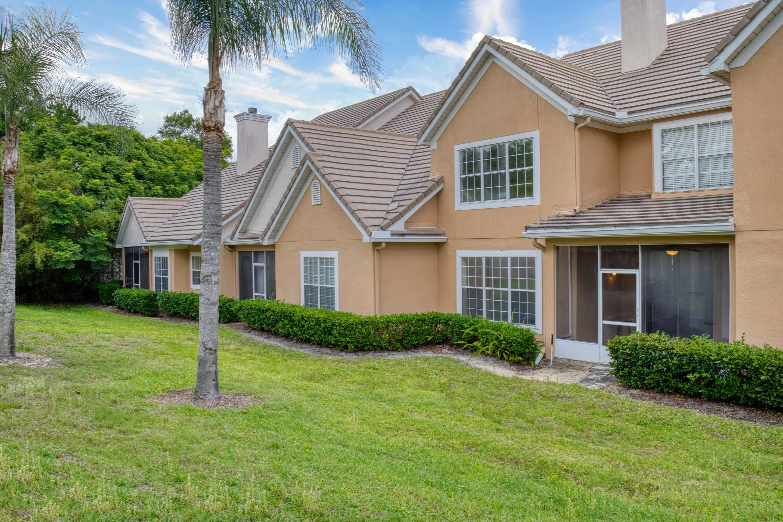 Multi-story beige house with large windows, surrounded by green grass, palm trees, and bushes, under a partly cloudy sky.