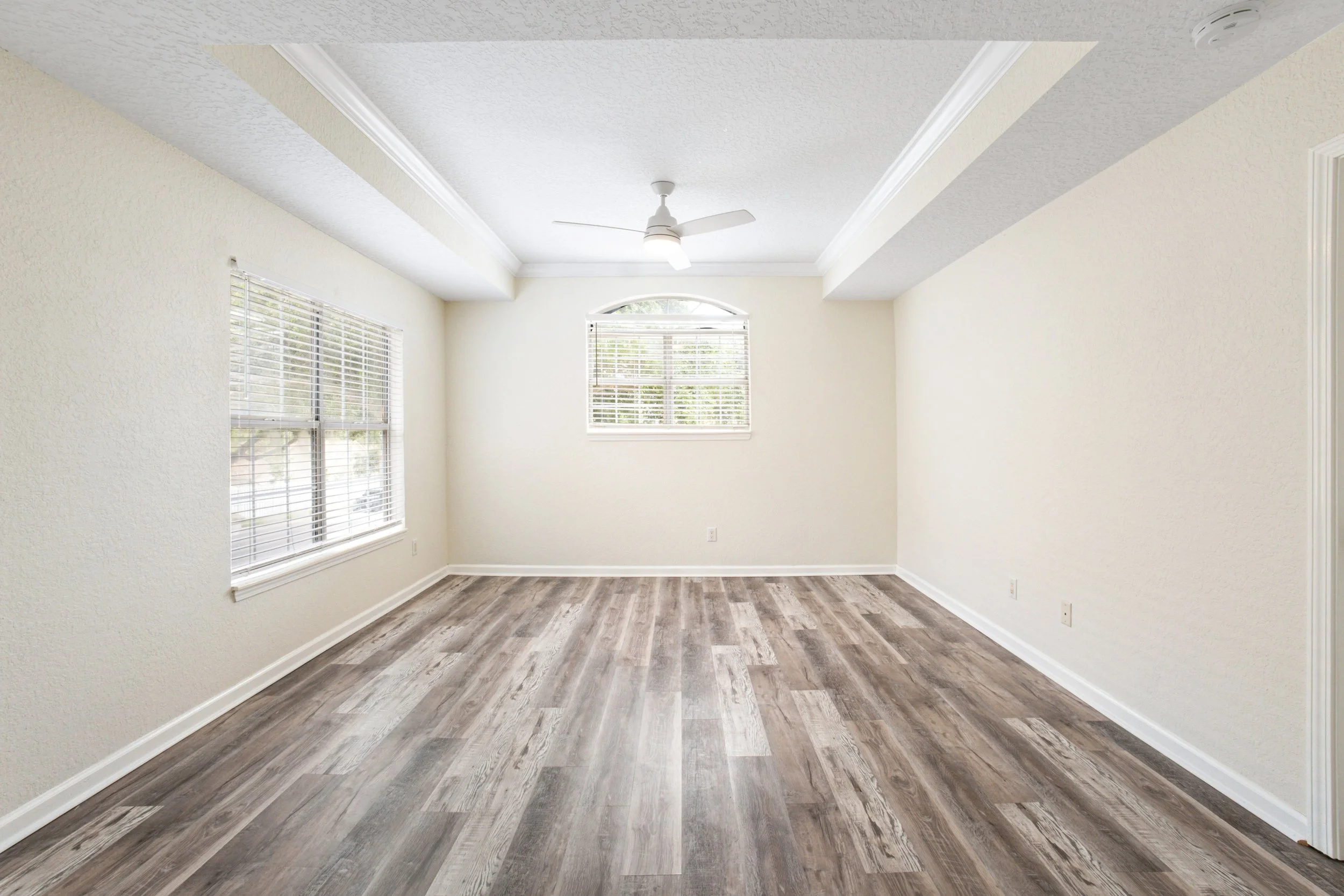Empty living room with two large windows, a ceiling fan, white walls, and wood-look flooring.