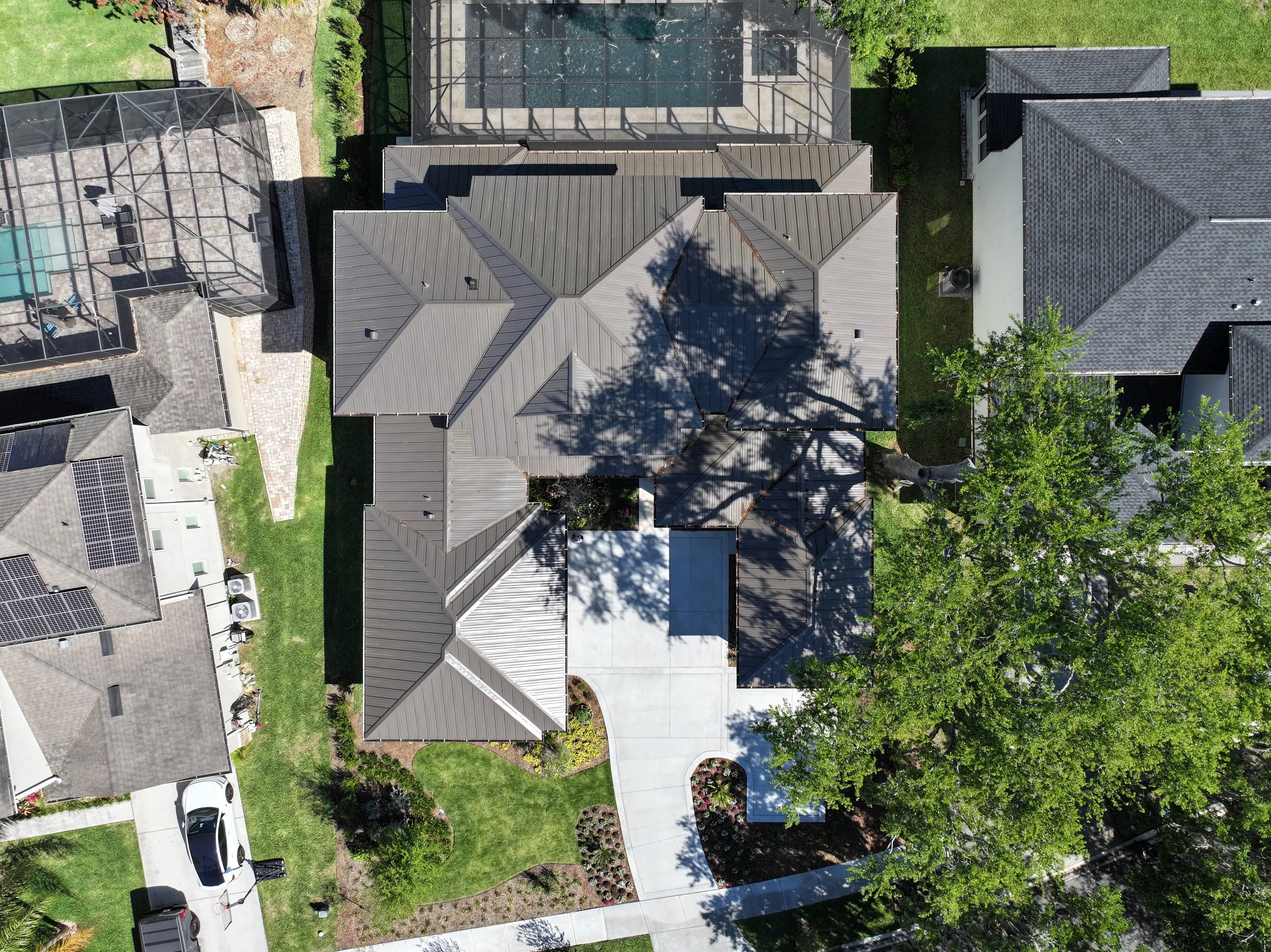 An aerial view of a residential backyard with a large house, a swimming pool enclosed in a screened structure, a patio area, and well-maintained lawns with trees and landscaping.