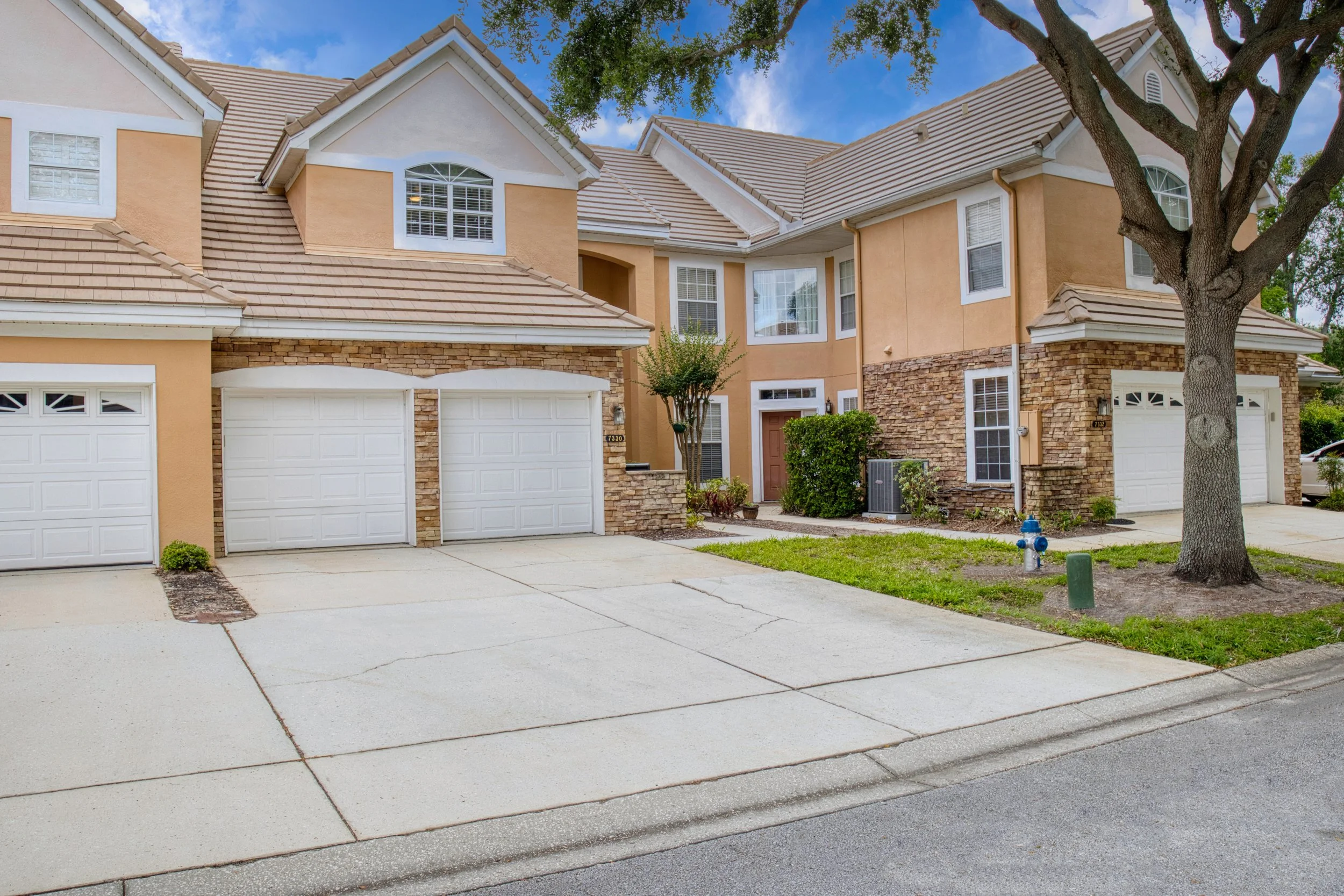A beige and brick two-story house with white trim, three garage doors, a driveway, a sidewalk, a tree, and some bushes in a suburban neighborhood under a blue sky.