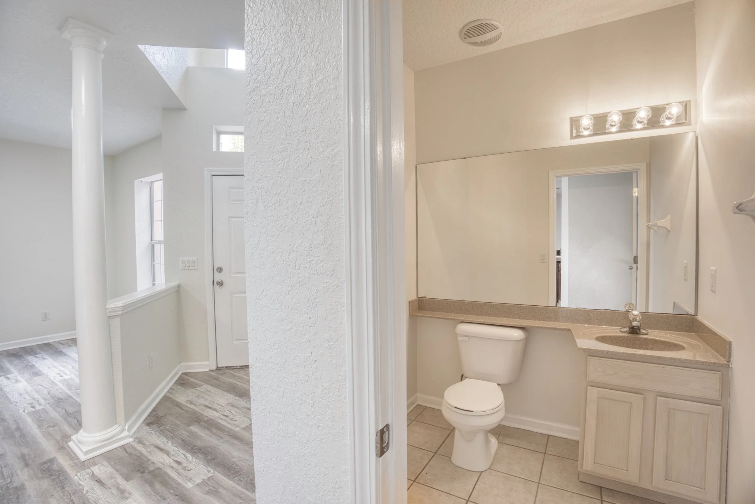 Bathroom with toilet, mirror, and sink, seen through a door frame. Adjacent room has white textured walls, a white column, and grey wood-look flooring.