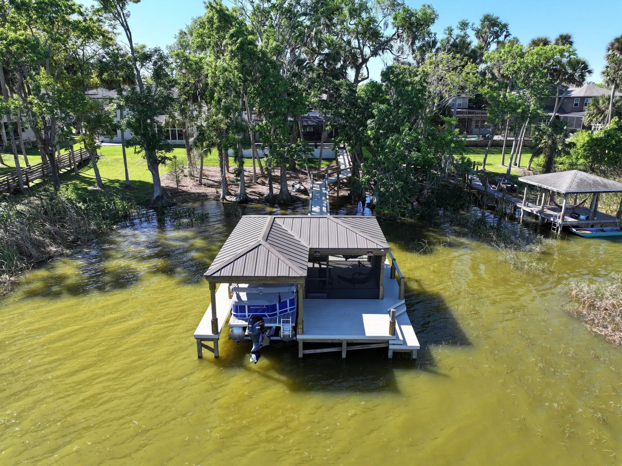 A river with a dock, a boat, and structures, surrounded by trees and houses in the background.