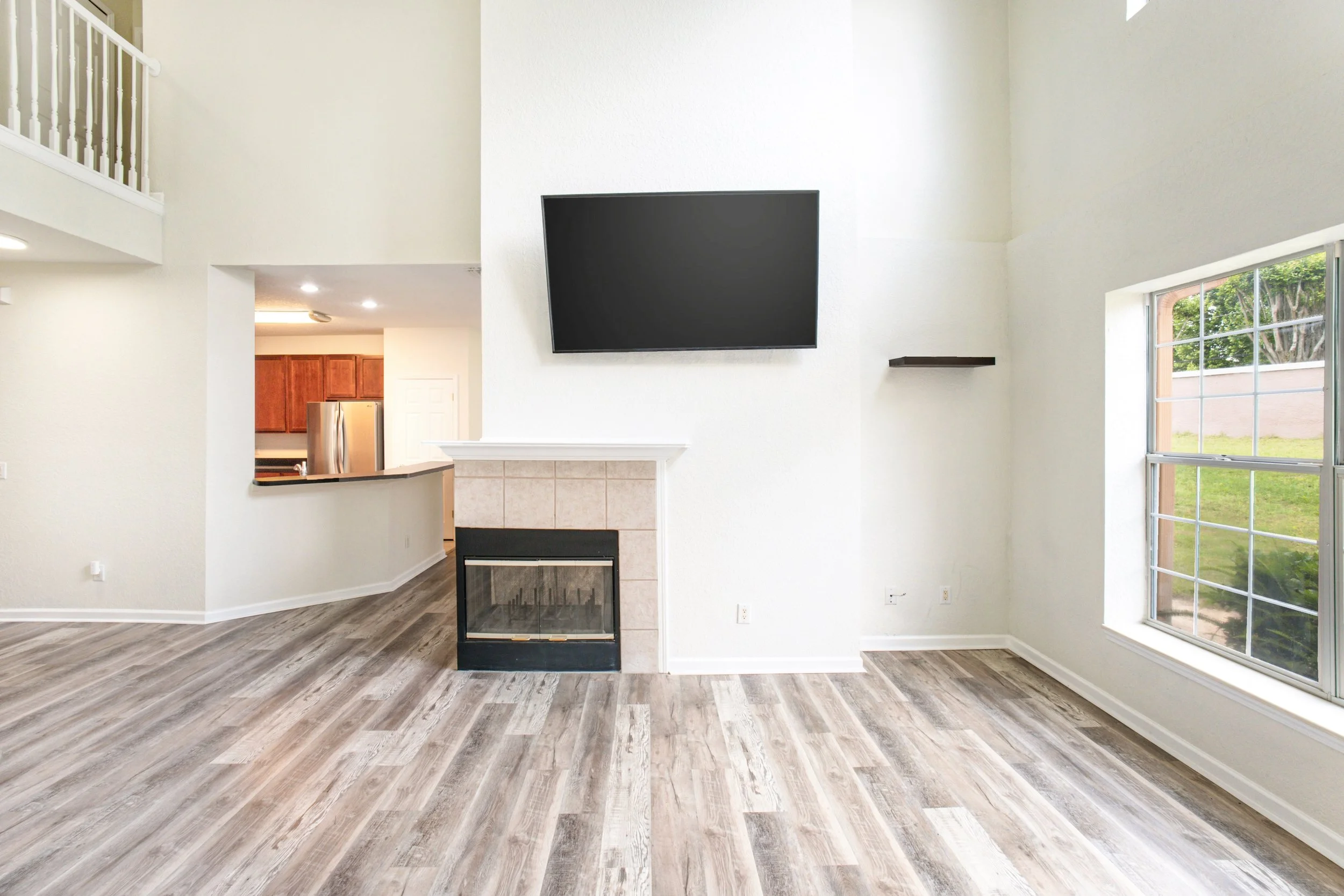 Interior of a modern living room with wood laminate flooring, a fireplace, a mounted flat-screen TV, large windows with a view of a backyard, and an open kitchen visible in the background.