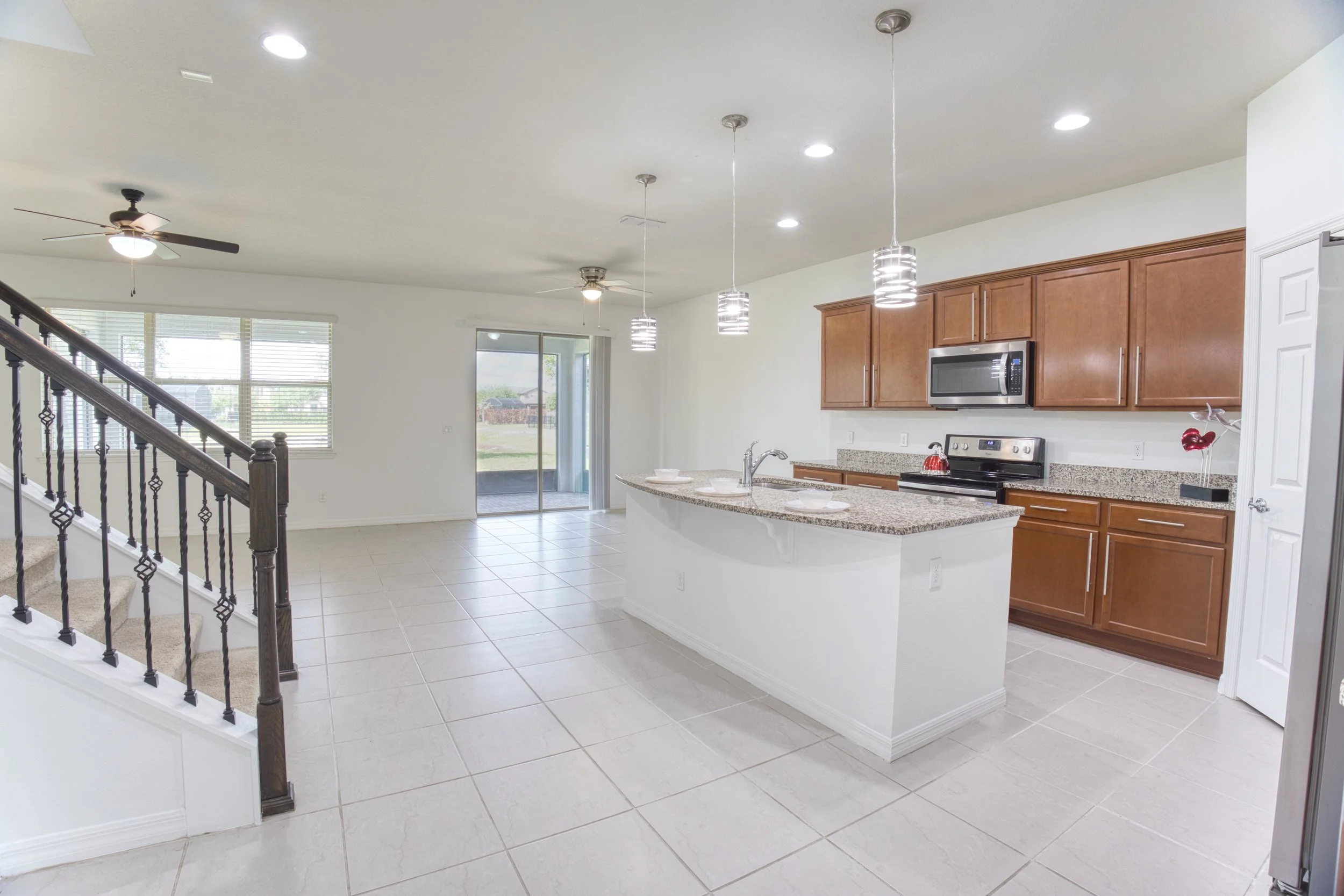 Open-concept kitchen with granite countertops, brown wooden cabinets, stainless steel microwave and stove, pendant lighting, ceiling fans, large window, sliding glass door, and tiled flooring.
