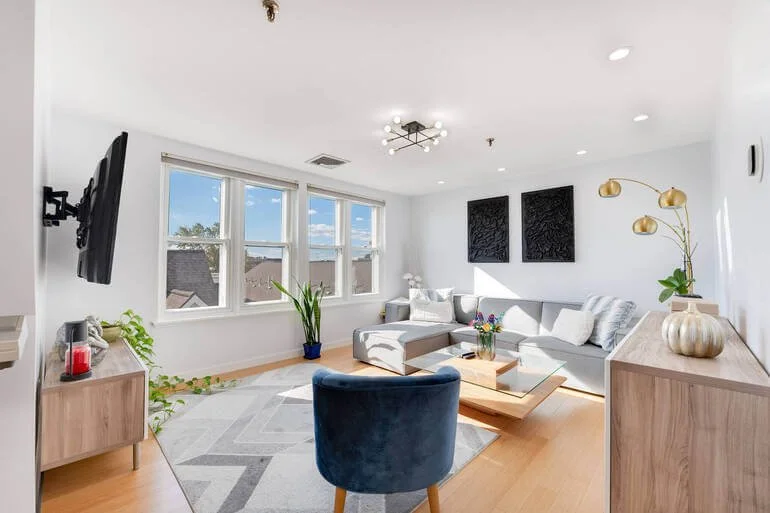 Bright living room with white walls, large window, gray sectional sofa, blue armchair, wooden coffee table, TV mounted on wall, and decorative plants and artwork.