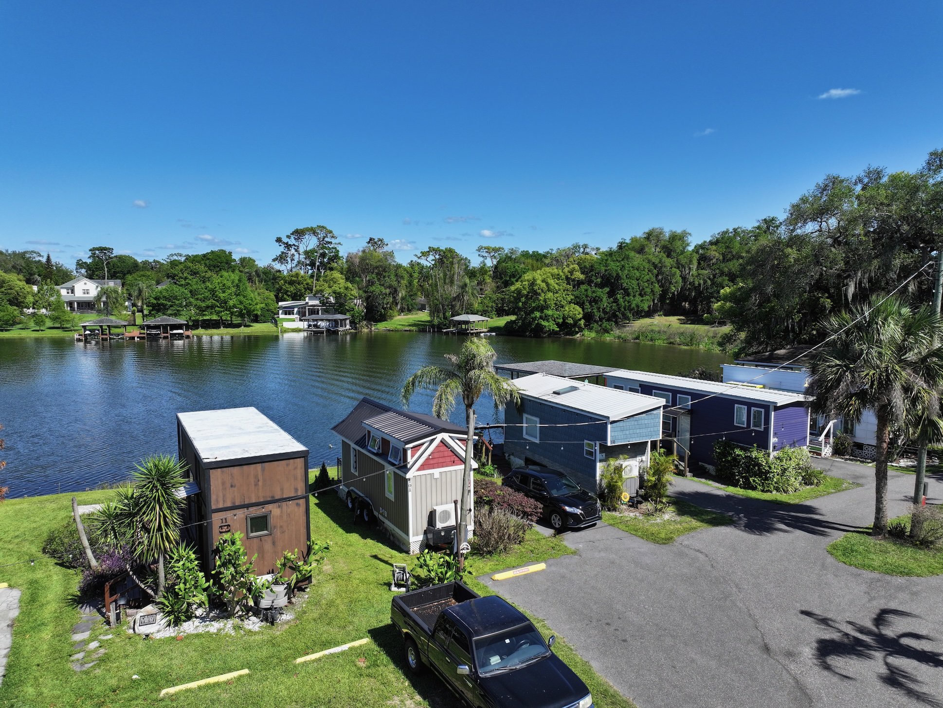 View of a lakeside neighborhood with small houses, trees, and a calm lake on a bright sunny day with a clear blue sky.