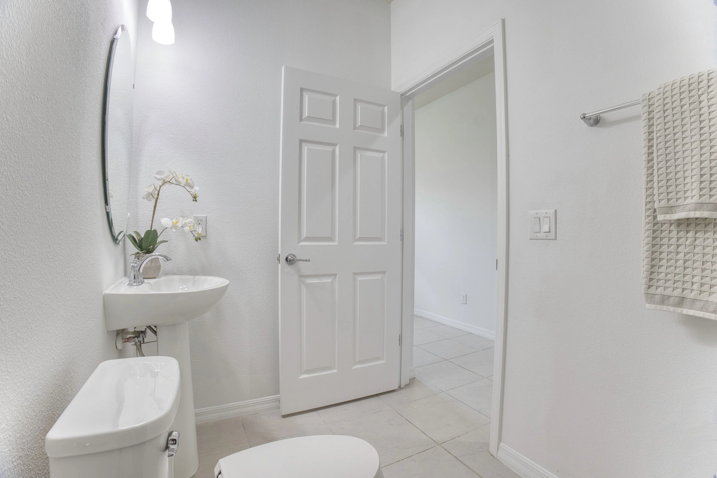 A bathroom with white walls and tile flooring, a small white sink with a silver faucet, a potted orchid plant on the sink, a mirror above, a closed white door, and a towel ring with a light-colored towel.