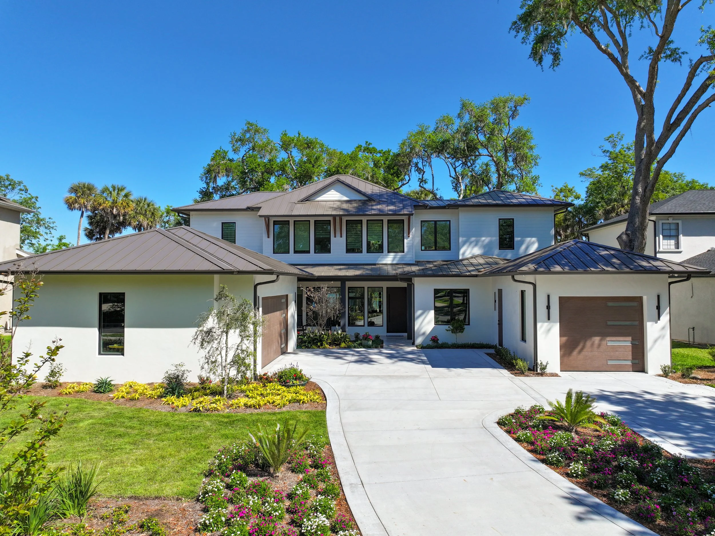Front view of a modern white house with large windows, surrounded by a well-maintained garden with small plants and colorful flowers, under a clear blue sky with tall trees in the background.