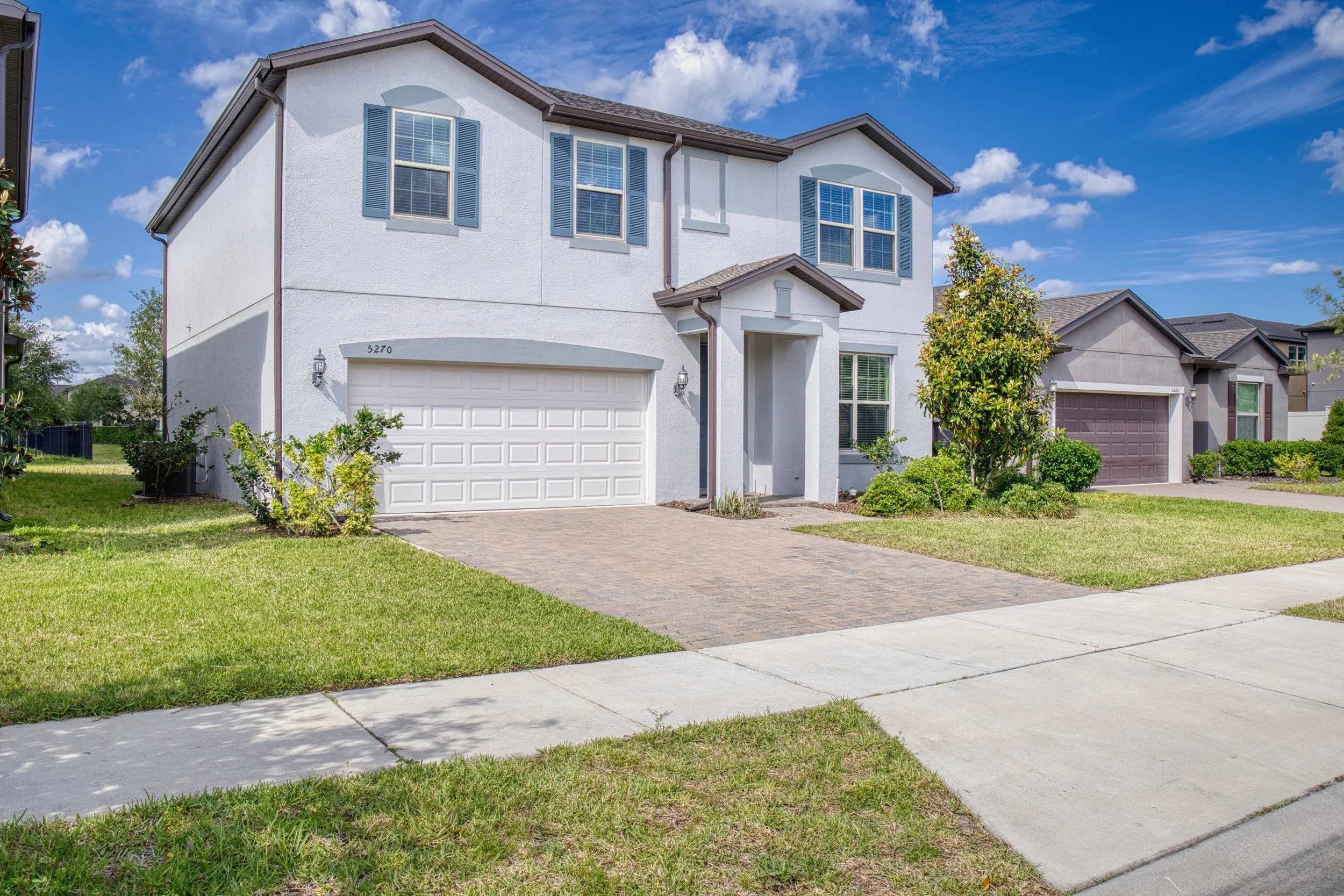 A two-story white suburban house with a brown roof, blue shutters, and a white garage door. The house has a small front porch, green lawn, and a brick-paved driveway. There are neighboring houses and a blue sky with scattered clouds.