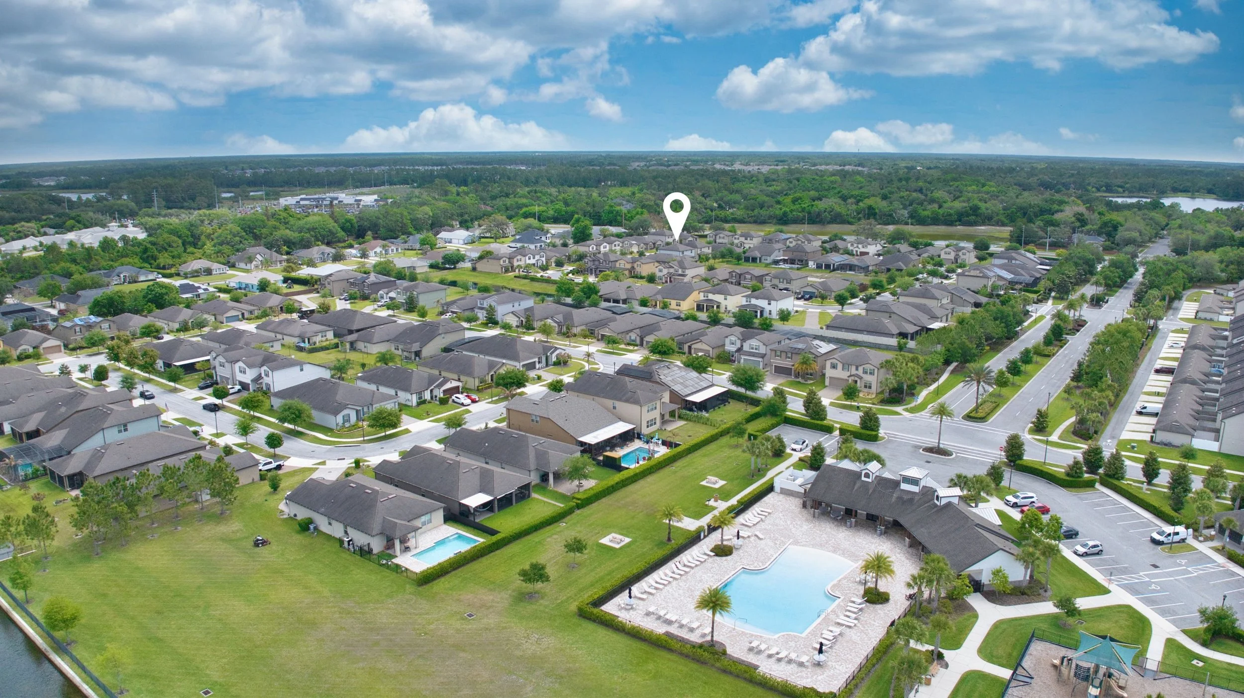 Aerial view of a residential neighborhood with houses, trees, and swimming pools, surrounded by greenery and a distant forest under a partly cloudy sky.