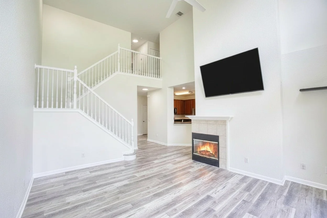 Empty living room with light-colored wood flooring, a fireplace, a wall-mounted television, and a staircase with white railings leading to an upper level.