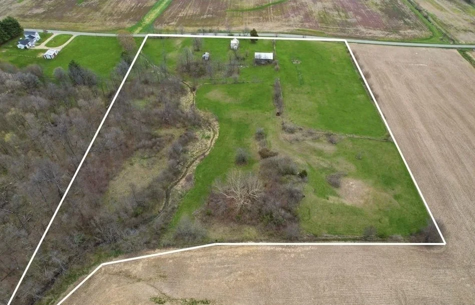 Aerial view of a large, irregularly shaped field with patches of trees, grass, and dirt, bordered by a white outline. Surrounding farmland and a few residential buildings are visible nearby.