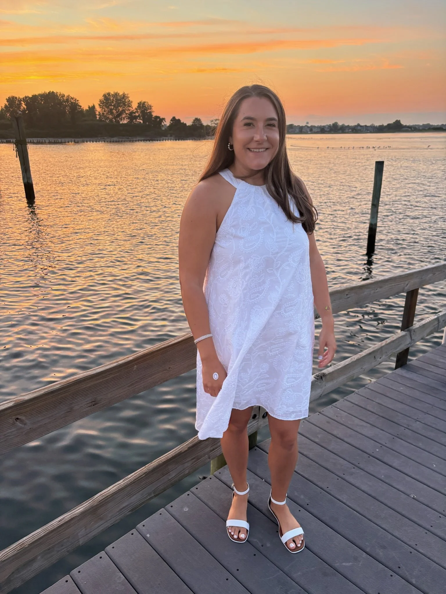 A woman smiling while standing on a wooden dock by a body of water during sunset, wearing a white sleeveless dress and white high-heeled sandals.