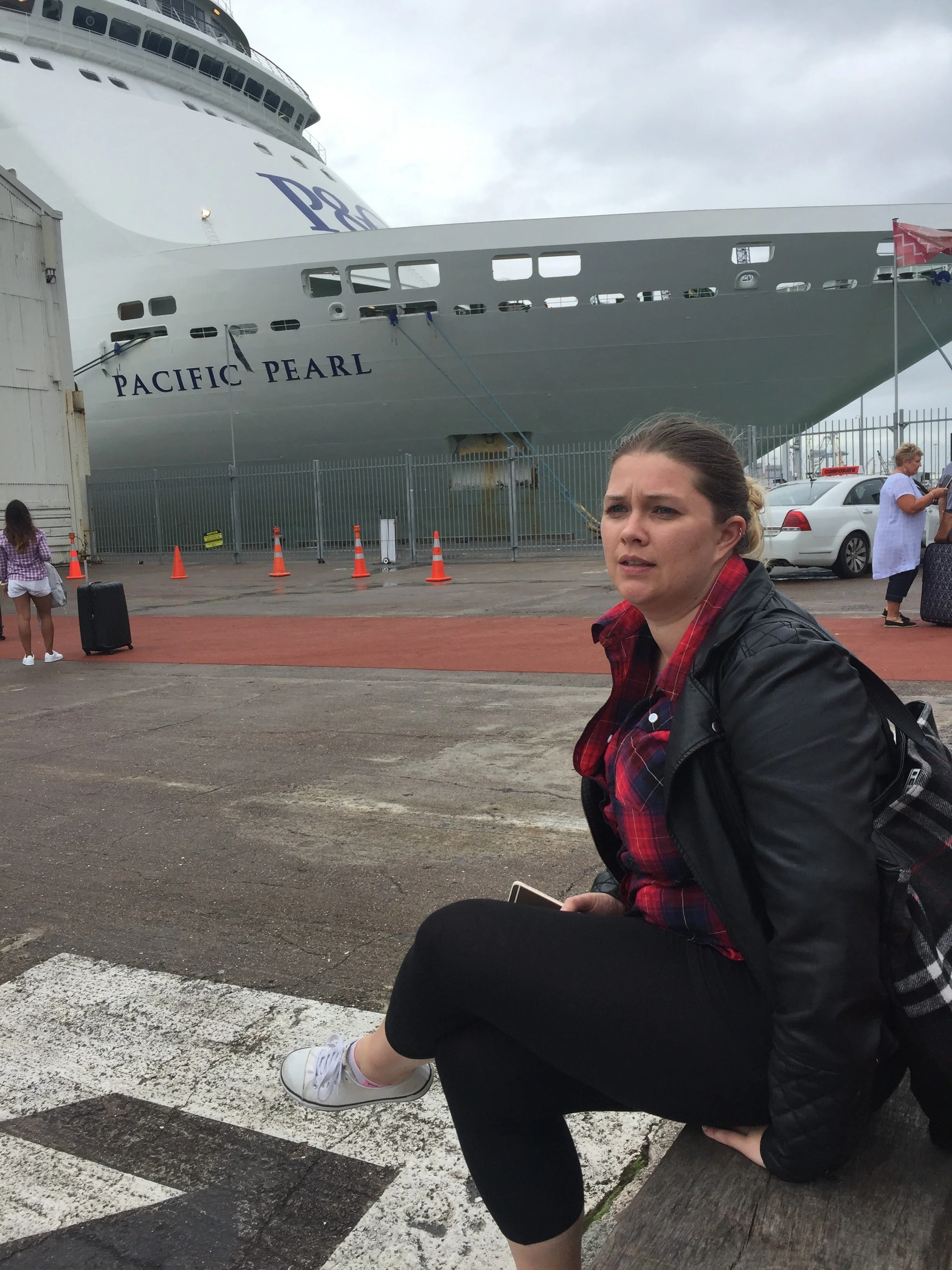 A woman sitting on a bench near a cruise ship named Pacific Pearl, with other people and cars nearby.