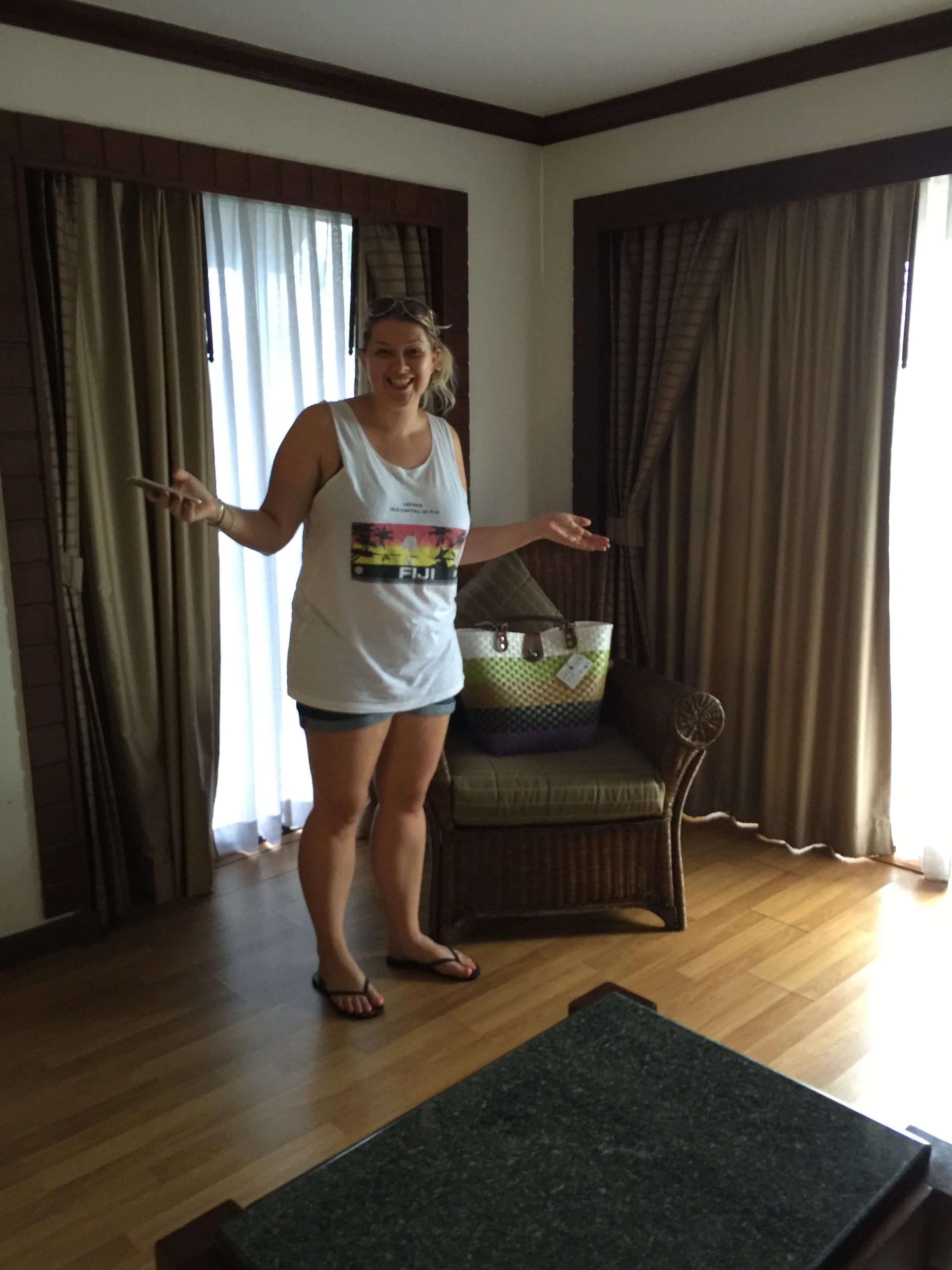 A woman with sunglasses on her head, wearing a white tank top and shorts, standing indoors near curtains, smiling and talking.