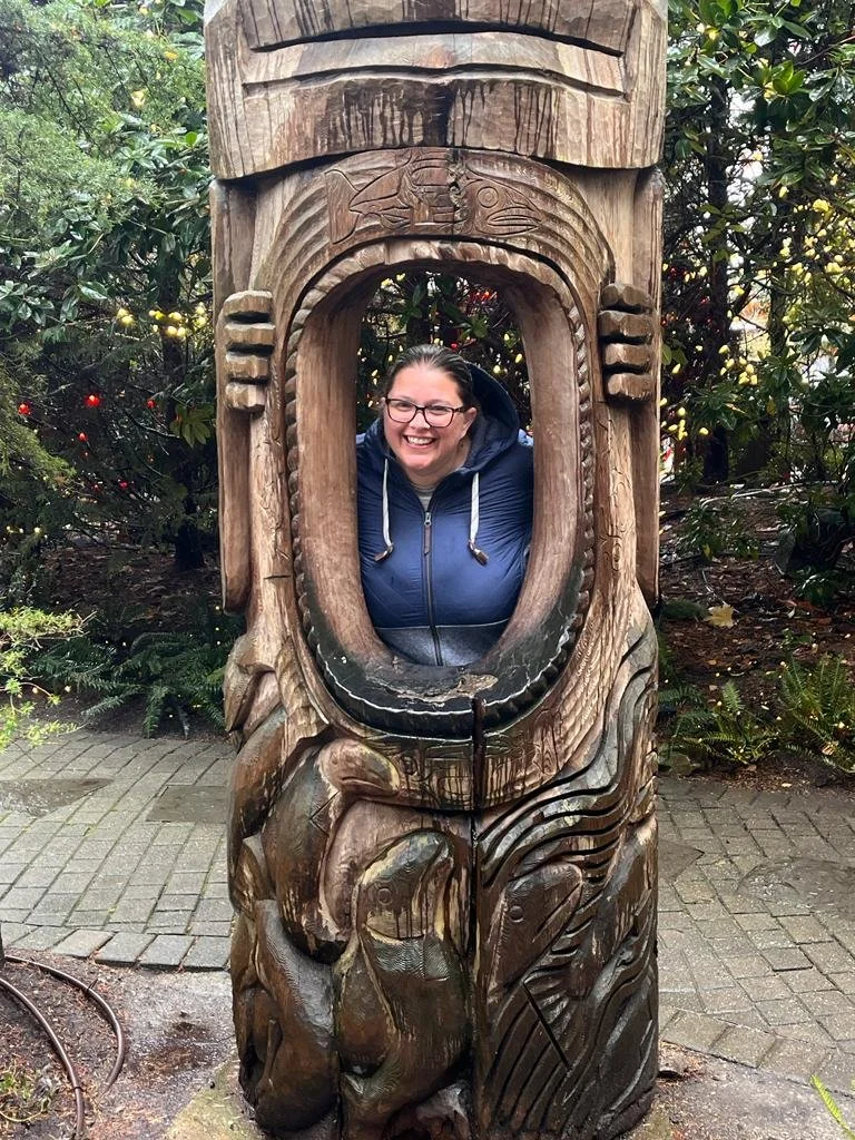 A woman with glasses and a blue jacket is smiling and posing behind a carved wooden figure with an open mouth, standing outdoors amidst green foliage.