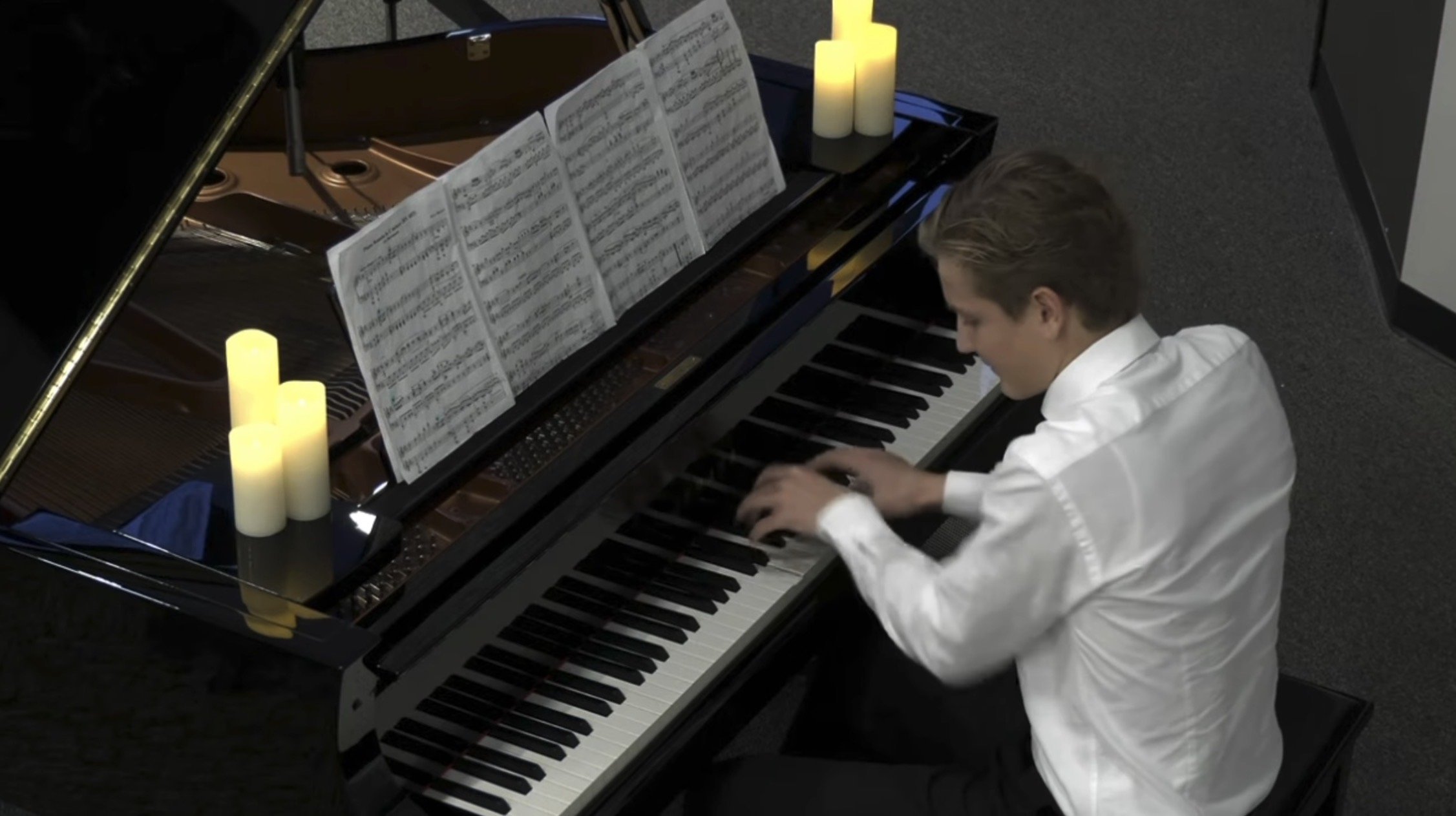 Young man playing a grand piano with sheet music and LED candles on top, in a dimly lit room.