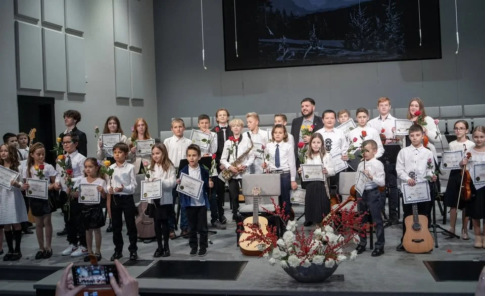 Children and adults on stage at an award ceremony, holding certificates and roses, with musical instruments and flowers in the foreground.