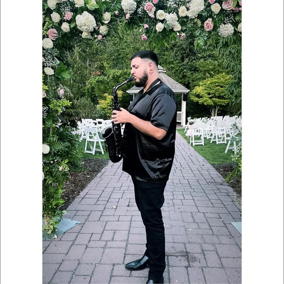 Man playing saxophone outdoors at a garden wedding ceremony with floral arch and white chairs.