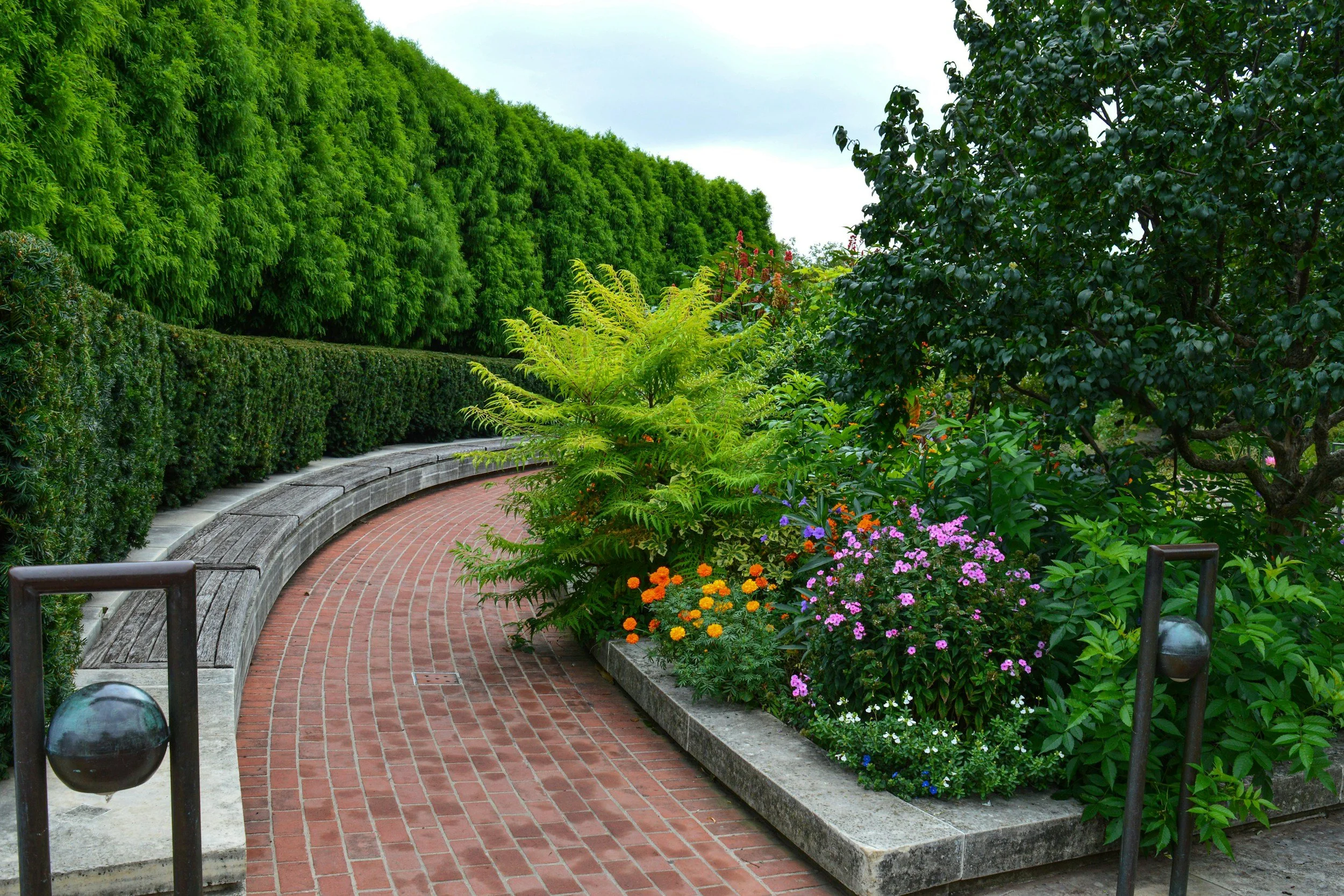 A curved brick pathway in a garden with green bushes and colorful flowers, surrounded by lush trees.