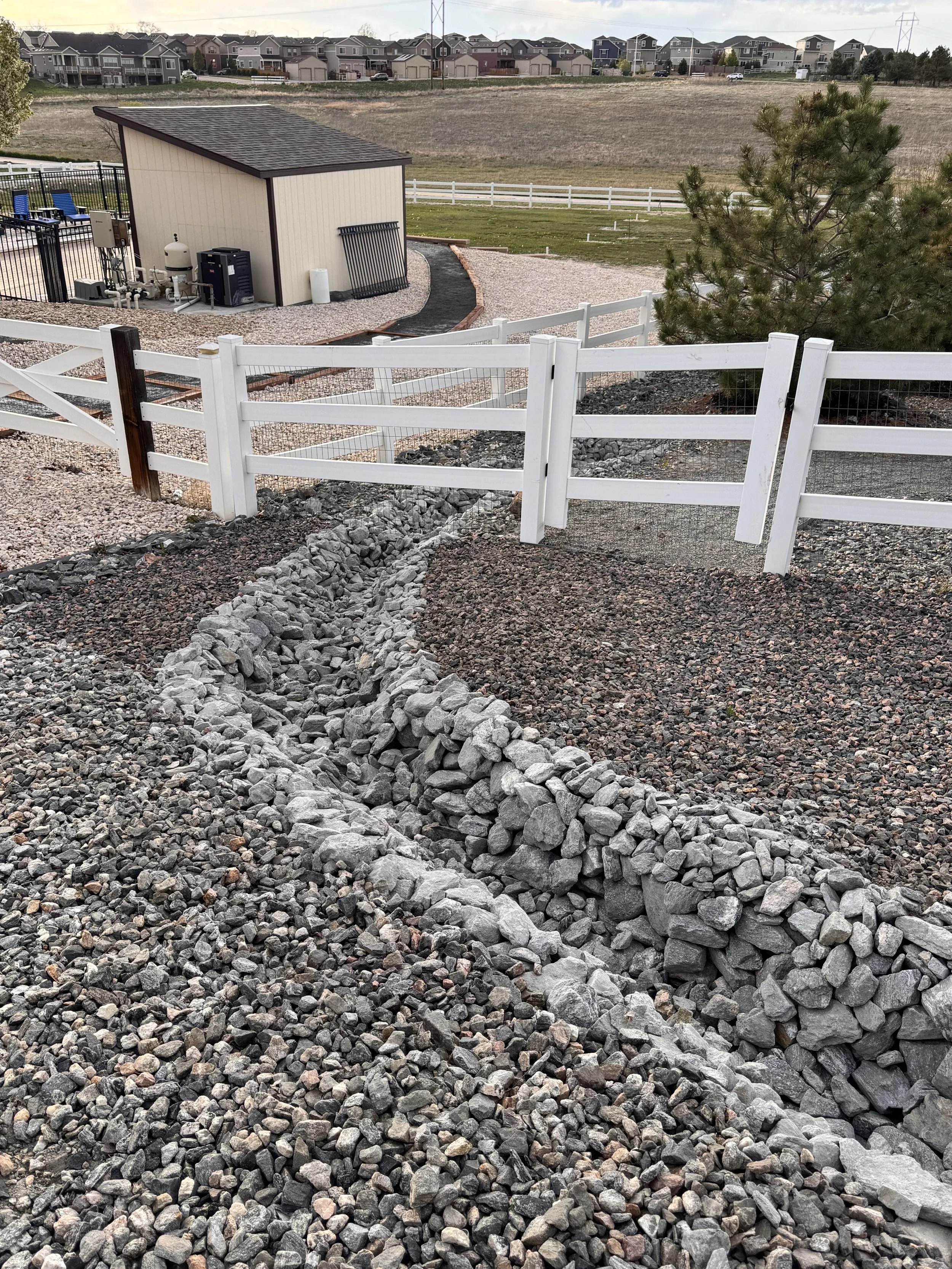 Stone-lined dry creek bed in a backyard with gravel and a white fence, a small shed, a pine tree, and a distant neighborhood of houses.