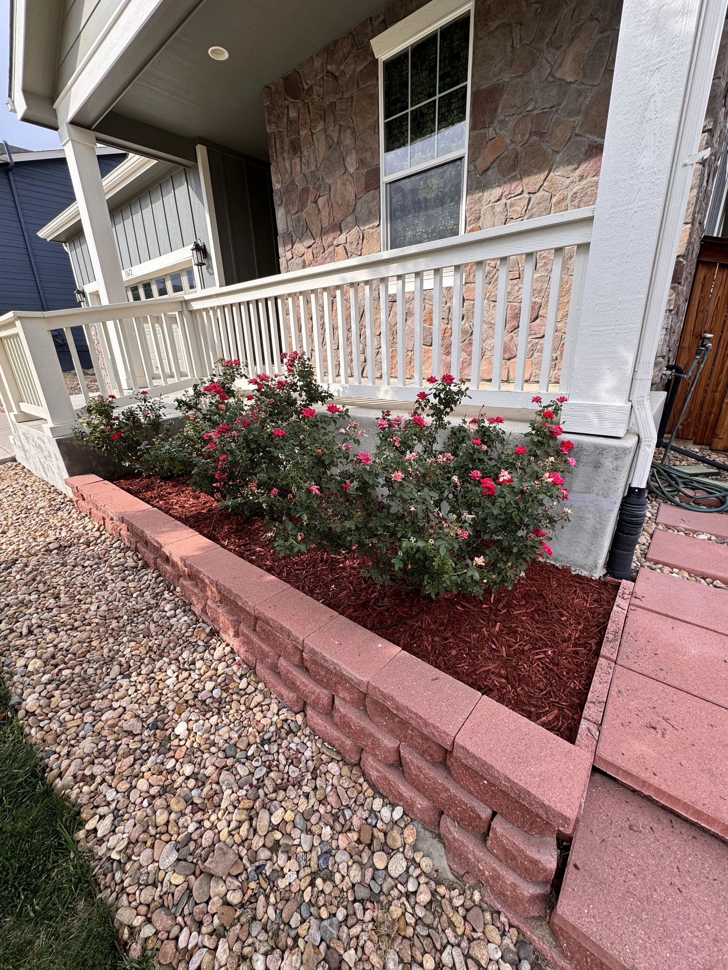 A house with a porch, white railing, a window, and a flower bed with pink and red roses, edged with red bricks, next to gray and pink paving stones and pebble ground.