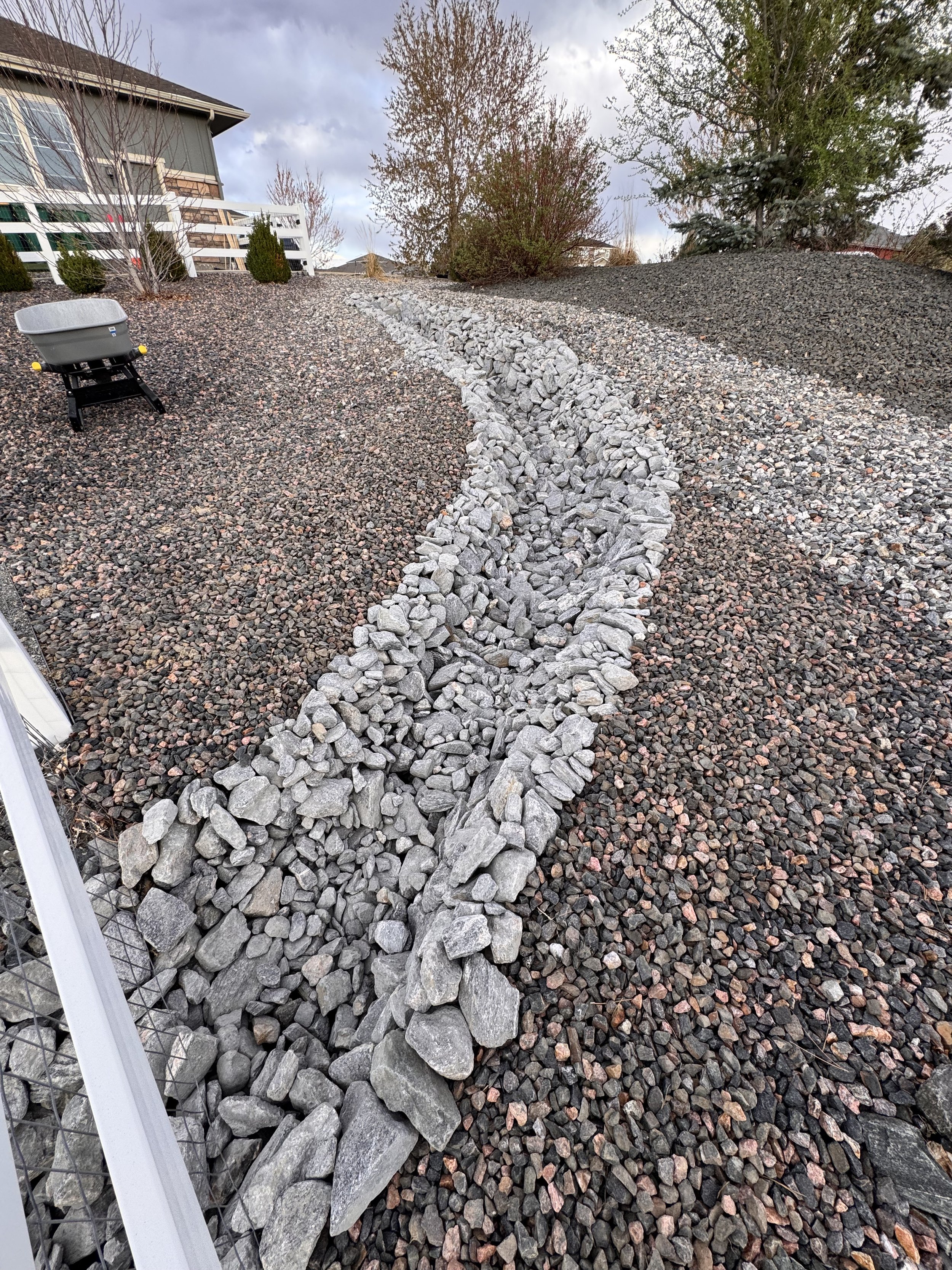 A landscaped yard with a gravel pathway edged with large gray rocks. The pathway curves up a slope towards the background, with a white fence, trees, and a house in the distance.