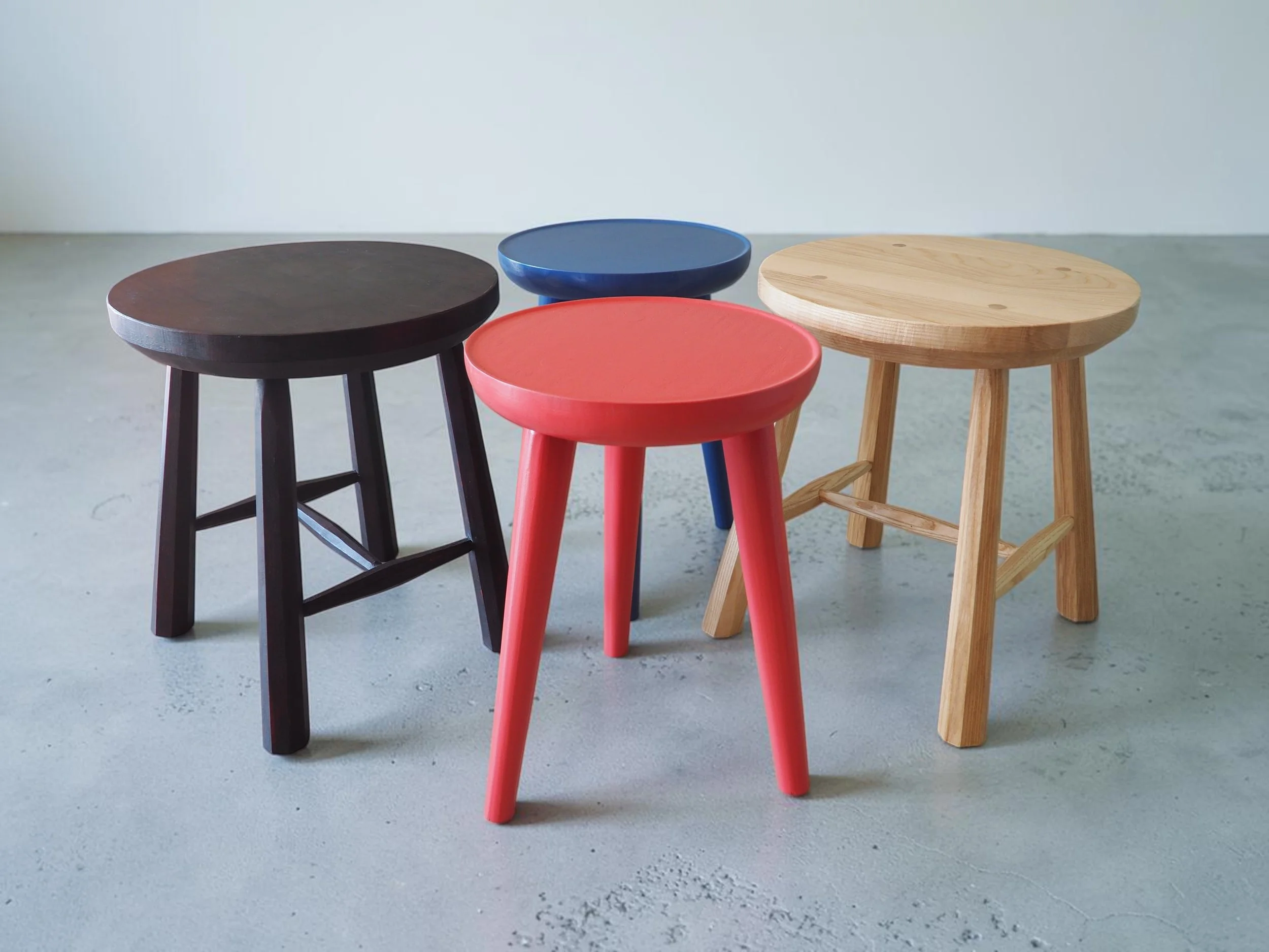 Five colorful wooden stools arranged in a semi-circle on a concrete floor against a plain light grey wall. The stools are in black, blue, red, natural wood, and dark brown colors.