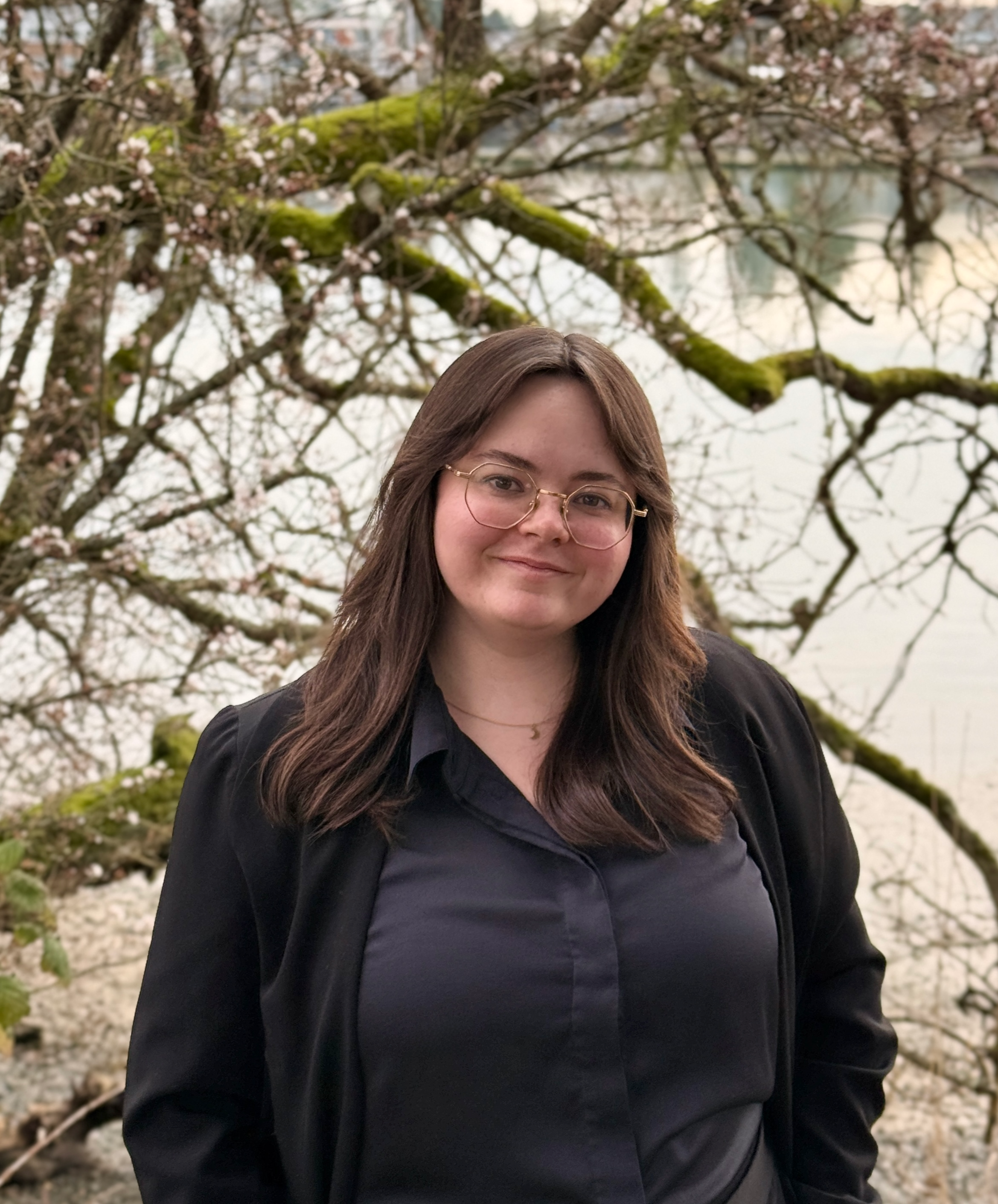 A woman with brown hair, glasses, and a black jacket standing outdoors in front of a tree with pink blossoms near a body of water.