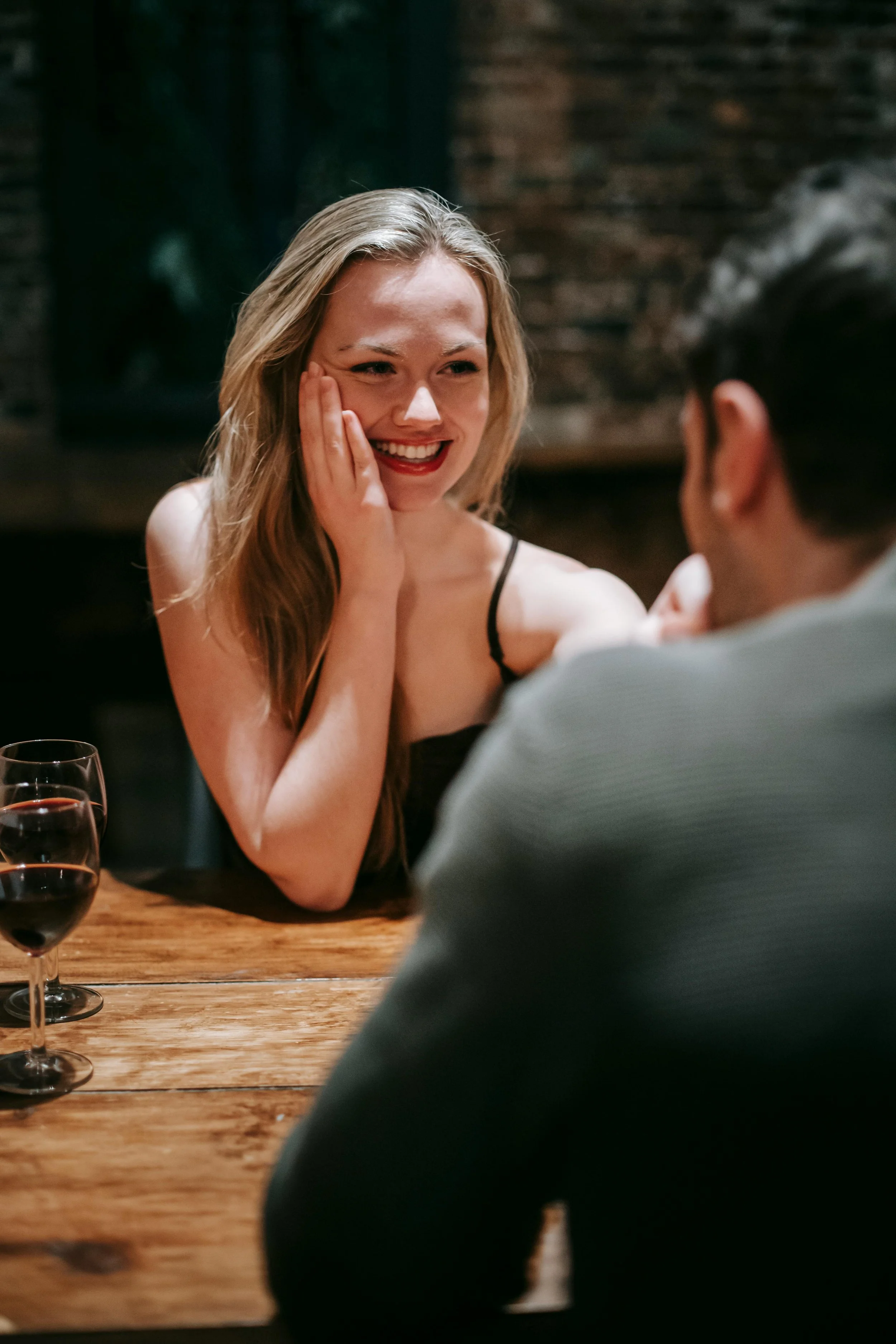 A woman with long blonde hair, smiling and touching her face, sitting at a wooden table with a man. There are two glasses of red wine on the table.