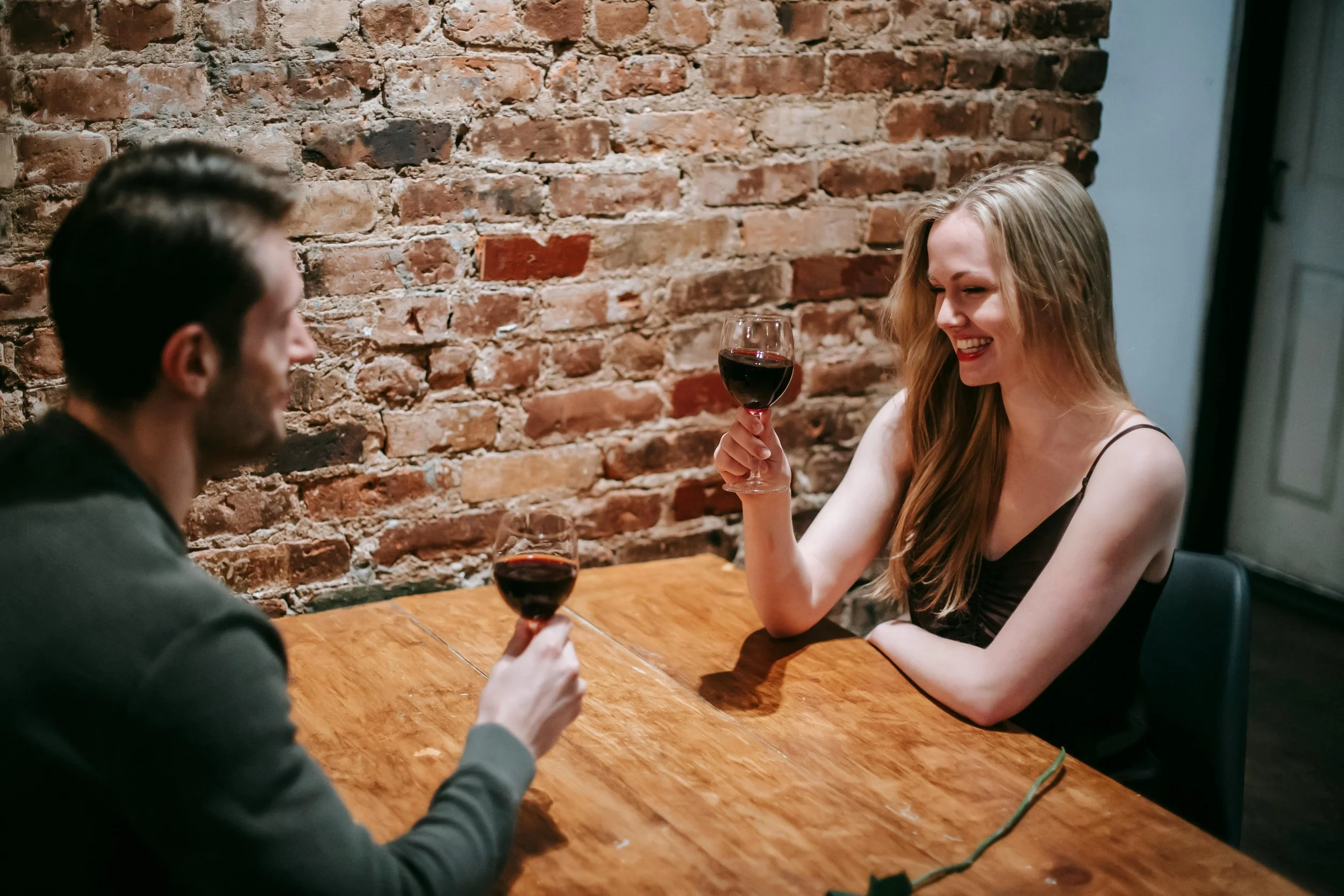 A man and woman sitting at a wooden table, drinking red wine and smiling at each other, with an exposed brick wall in the background.