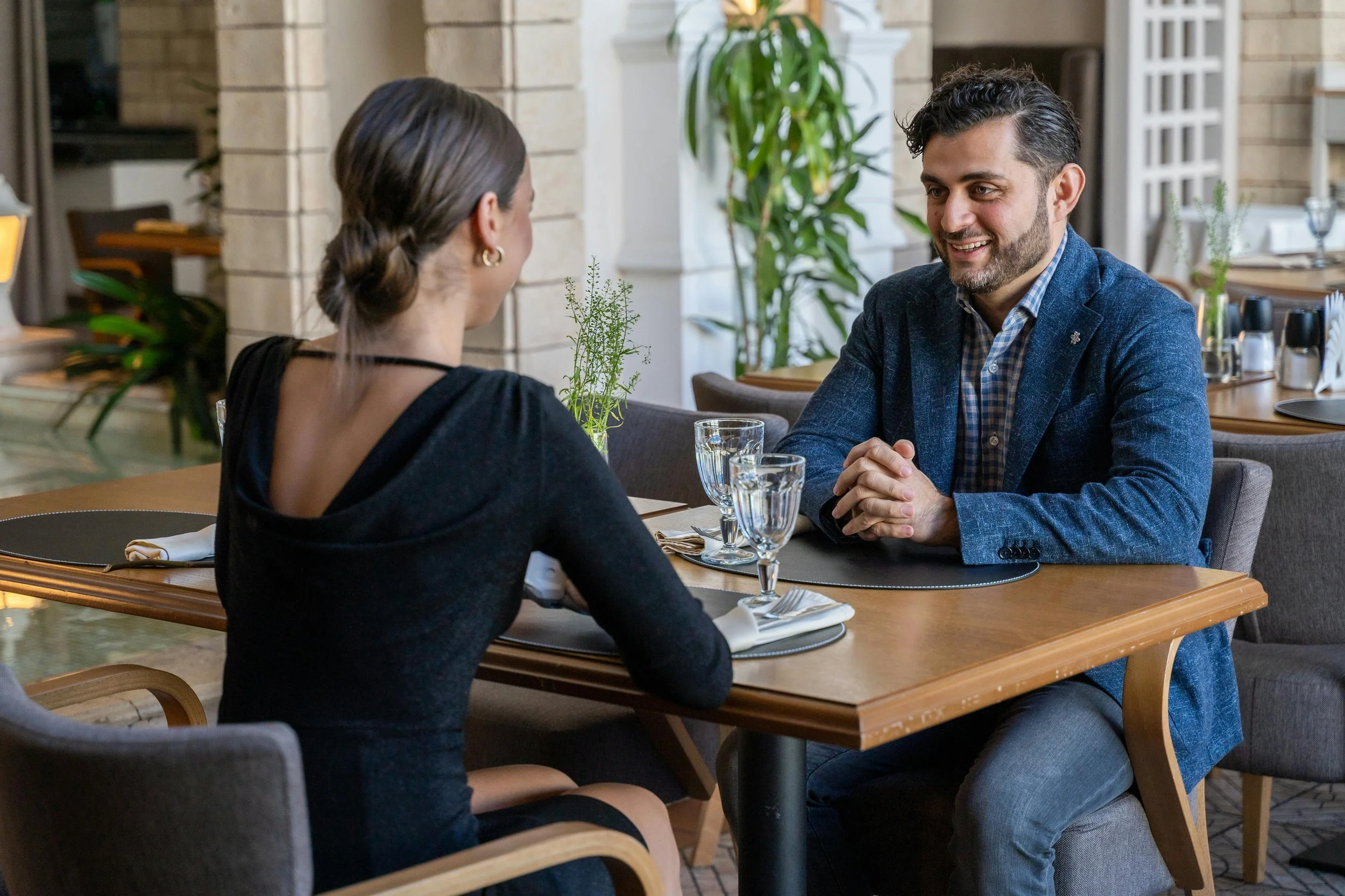 A man and woman having a conversation at a restaurant table, smiling and making eye contact, with water glasses and place settings on the table.