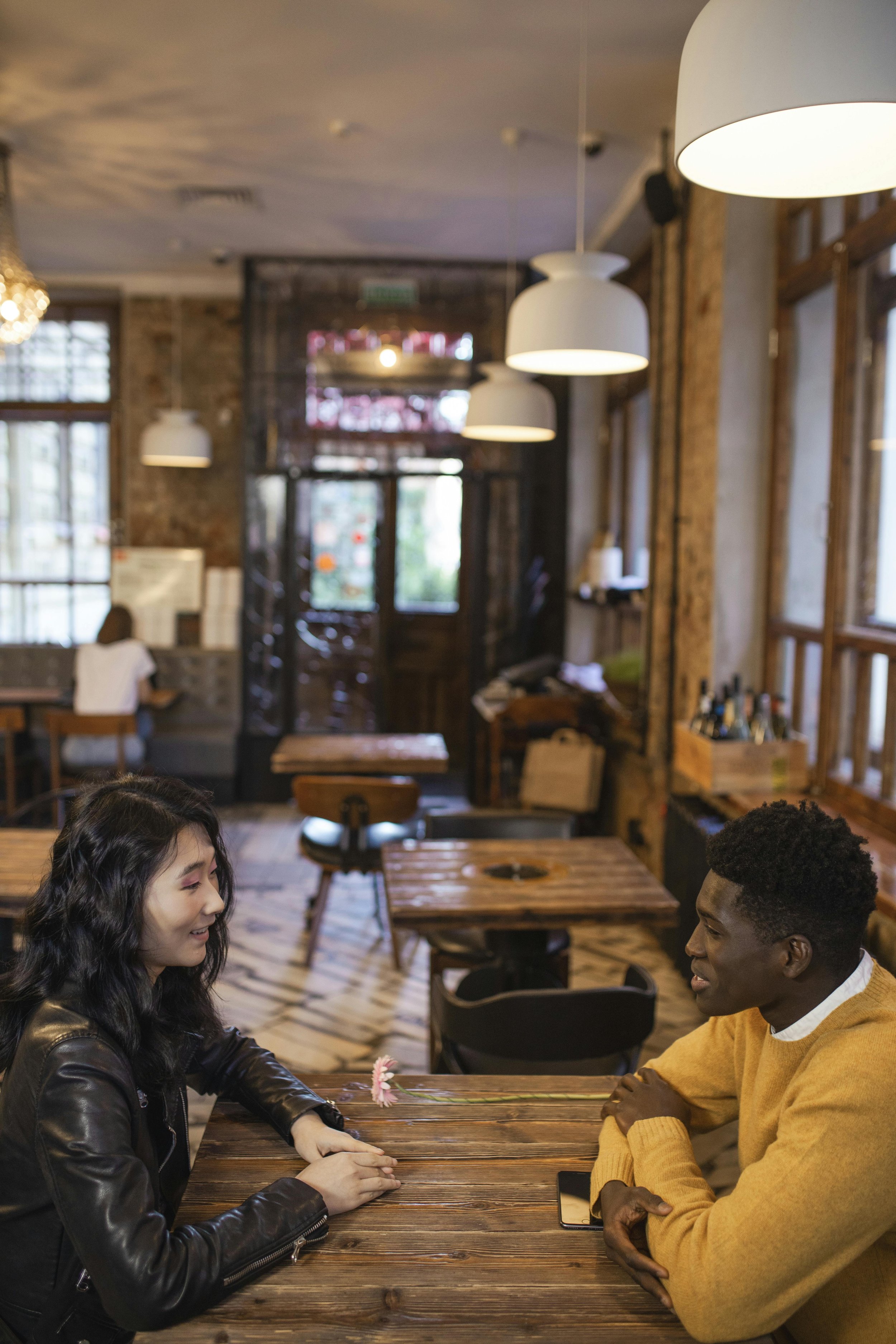 A woman and a man sitting across from each other at a wooden table in a cozy, rustic cafe, smiling and talking. The cafe has warm lighting, wooden decor, and large windows.