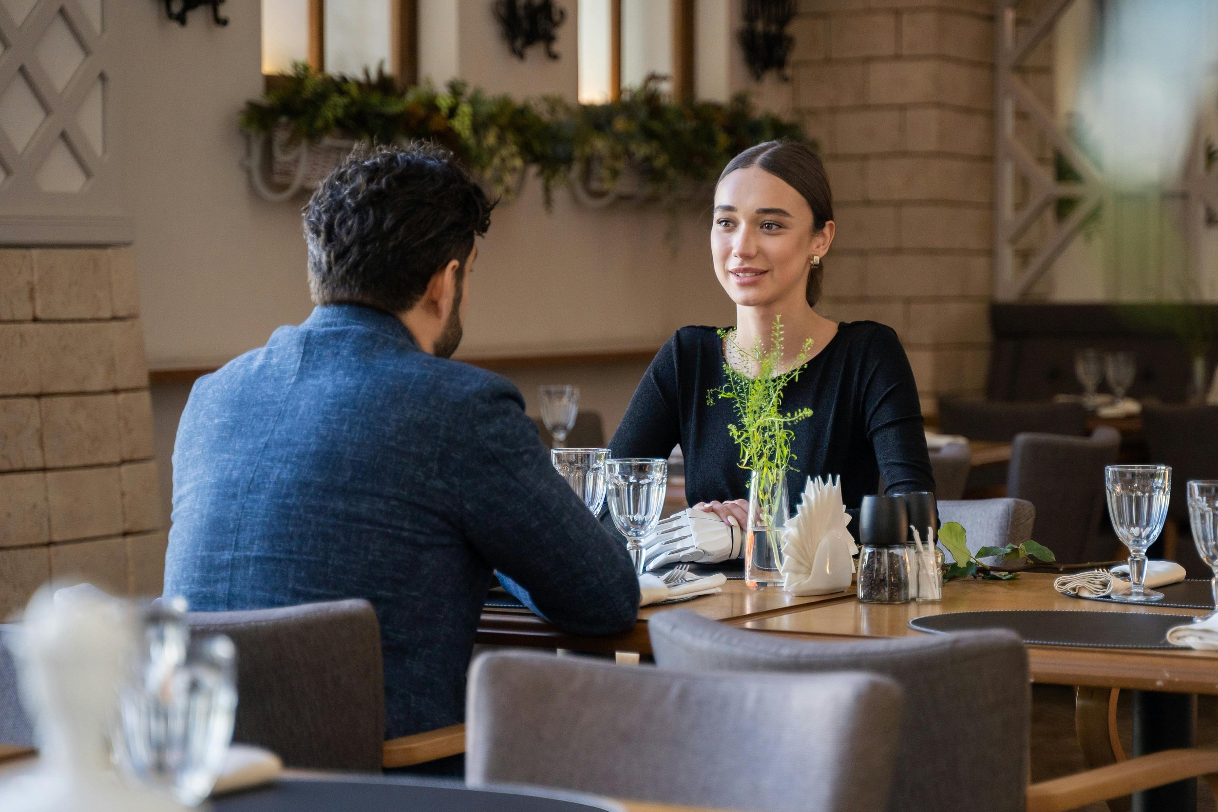 A man and woman having a conversation at a restaurant table, with wine glasses, a small vase with green plants, and neatly folded napkins.