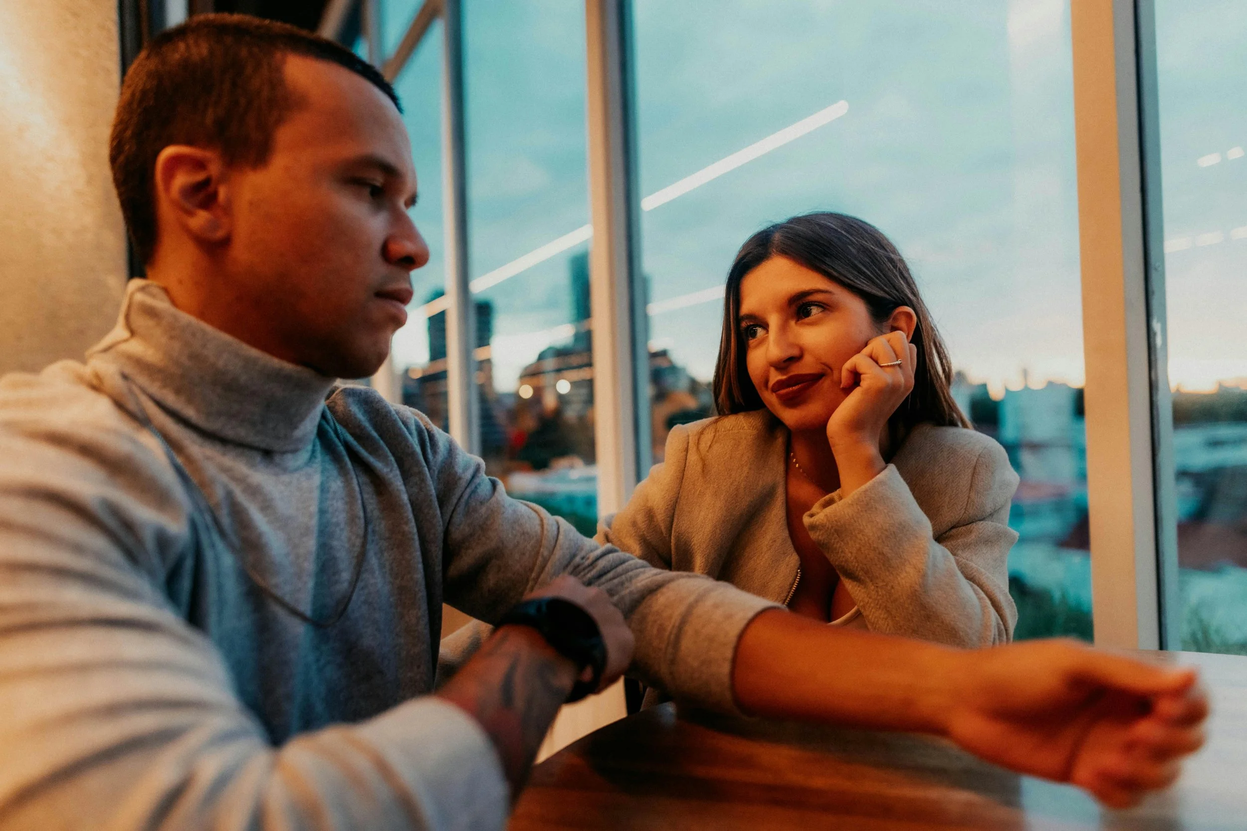 A young man and woman sitting at a wooden table near large windows, having a conversation in a restaurant or cafe during sunset.