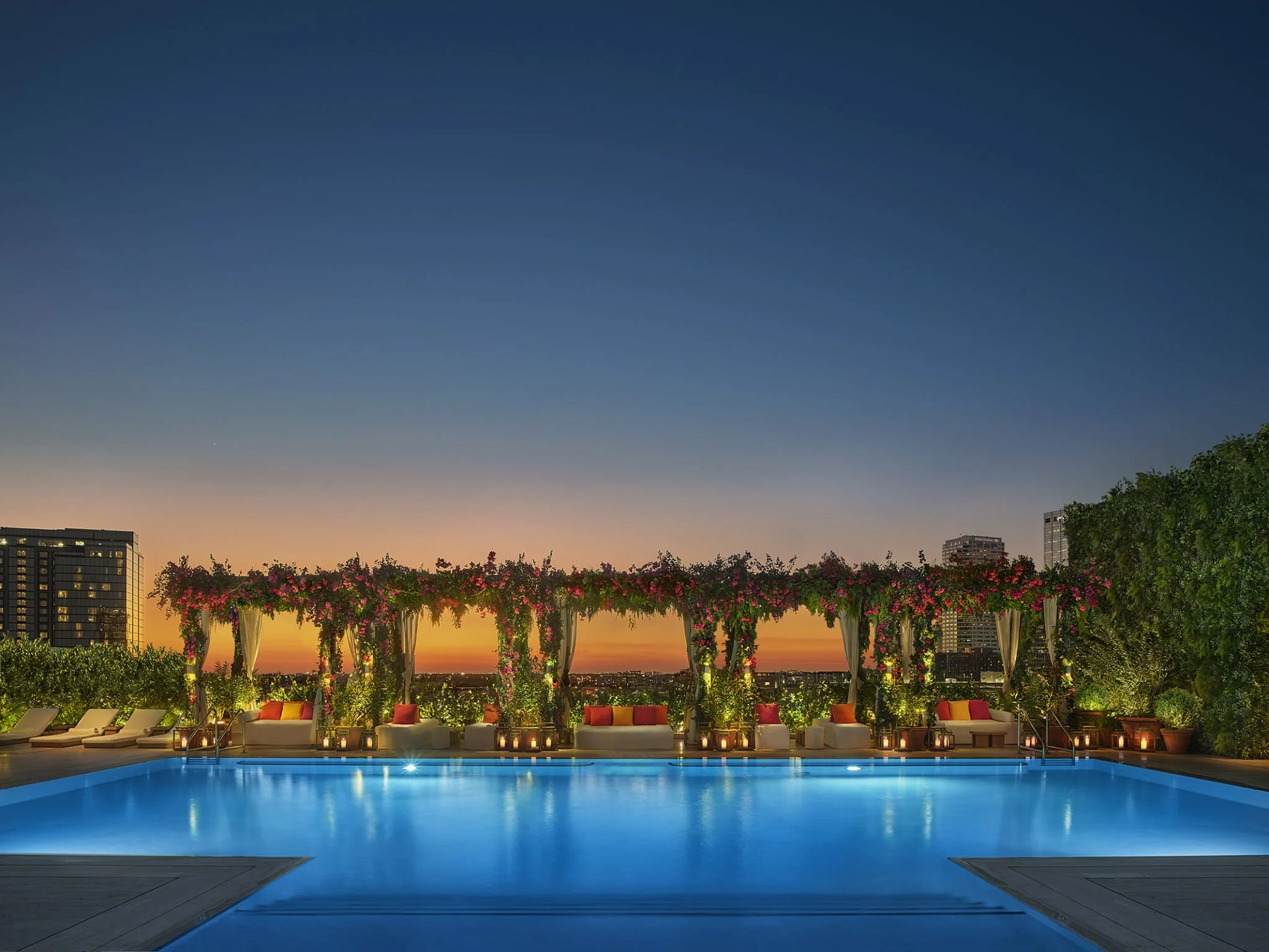 Rooftop swimming pool at sunset with floral cabana, city skyline, and lounge chairs.