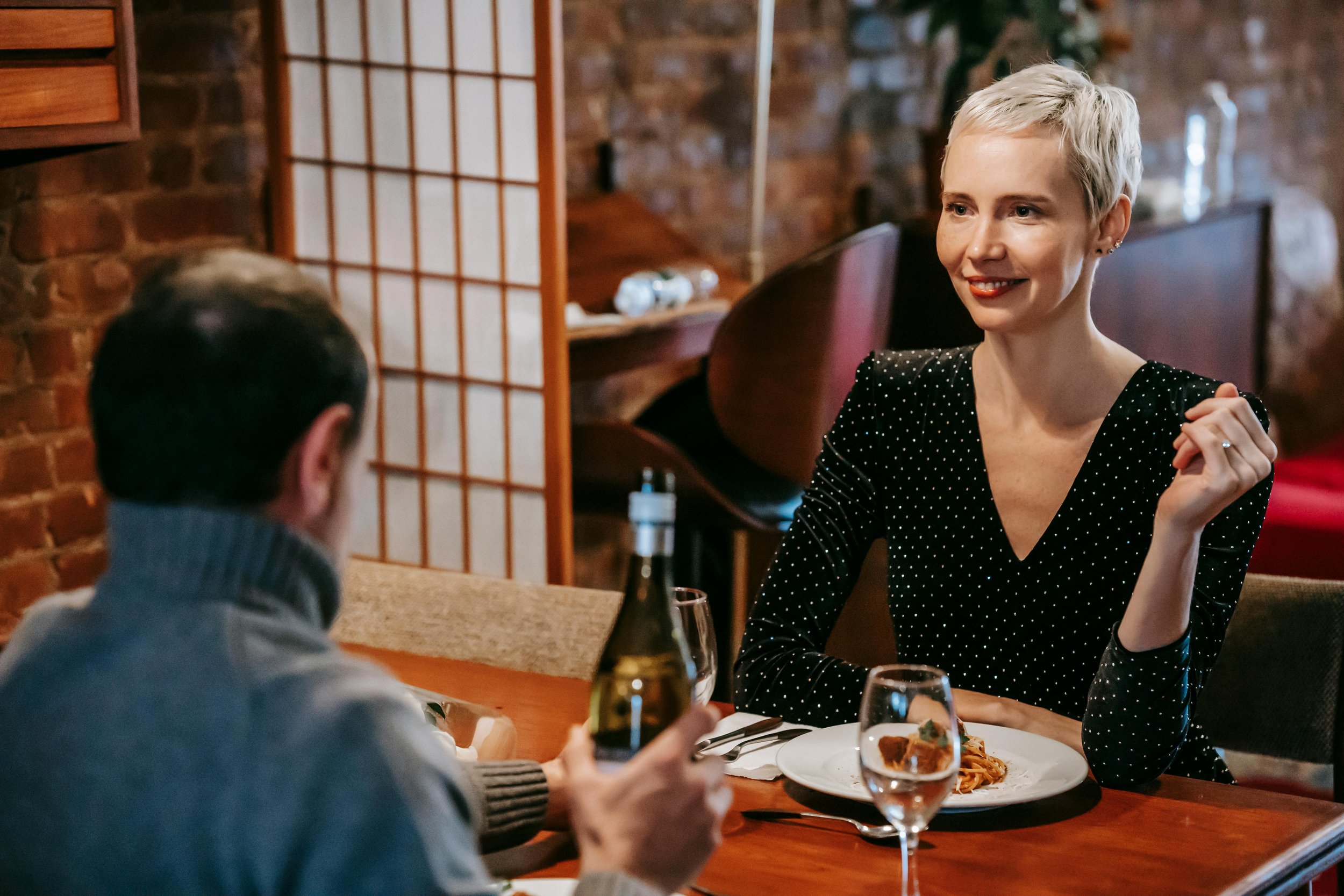 A woman with short blonde hair wearing a black polka dot dress is sitting at a restaurant table, smiling and looking at a man with dark hair holding a beer bottle. The table has a plate of spaghetti, a wine glass, and a fork. The restaurant has brick walls and wooden furniture.