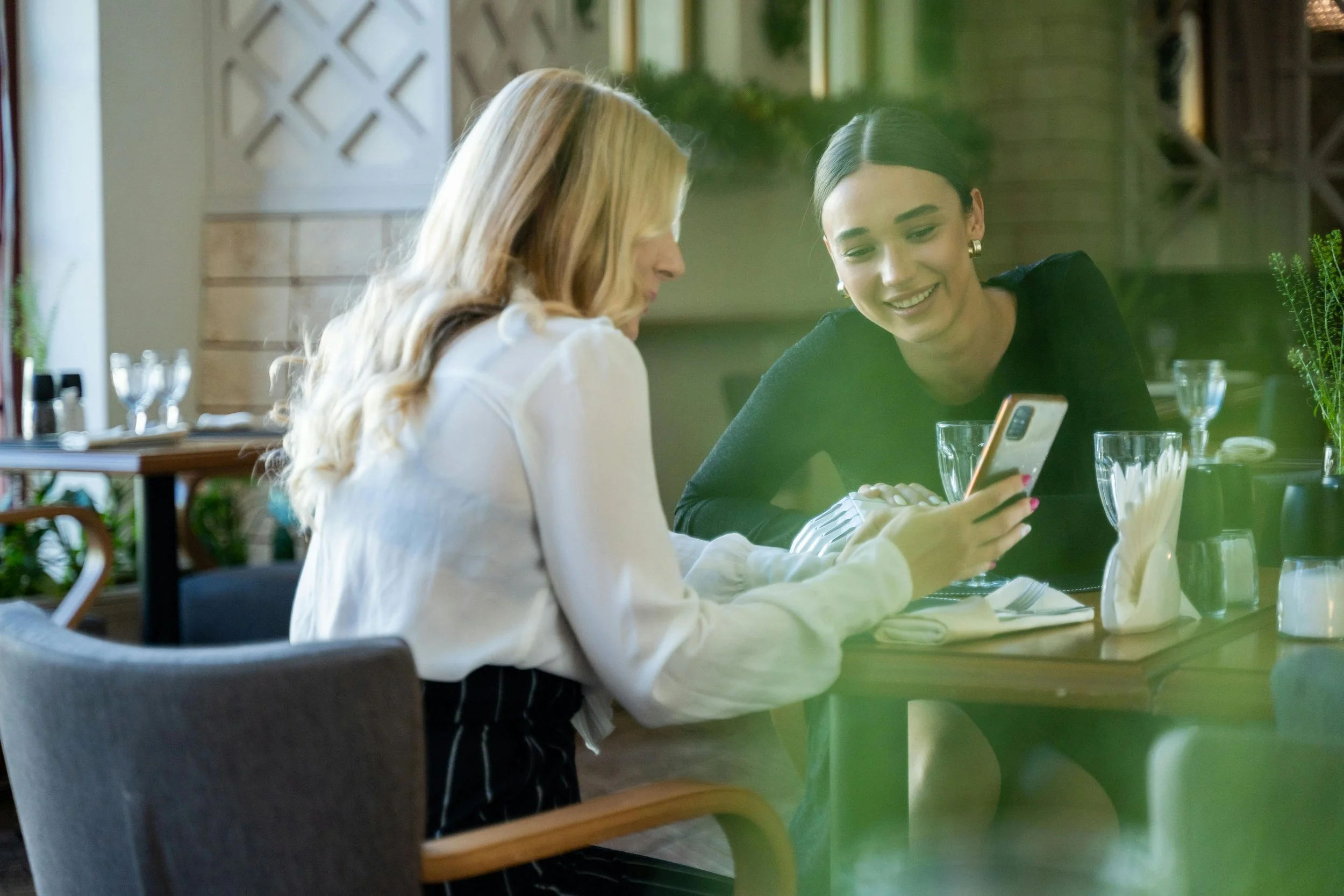 Two women sitting at a restaurant table, looking at a smartphone, smiling, with glasses and table settings visible.